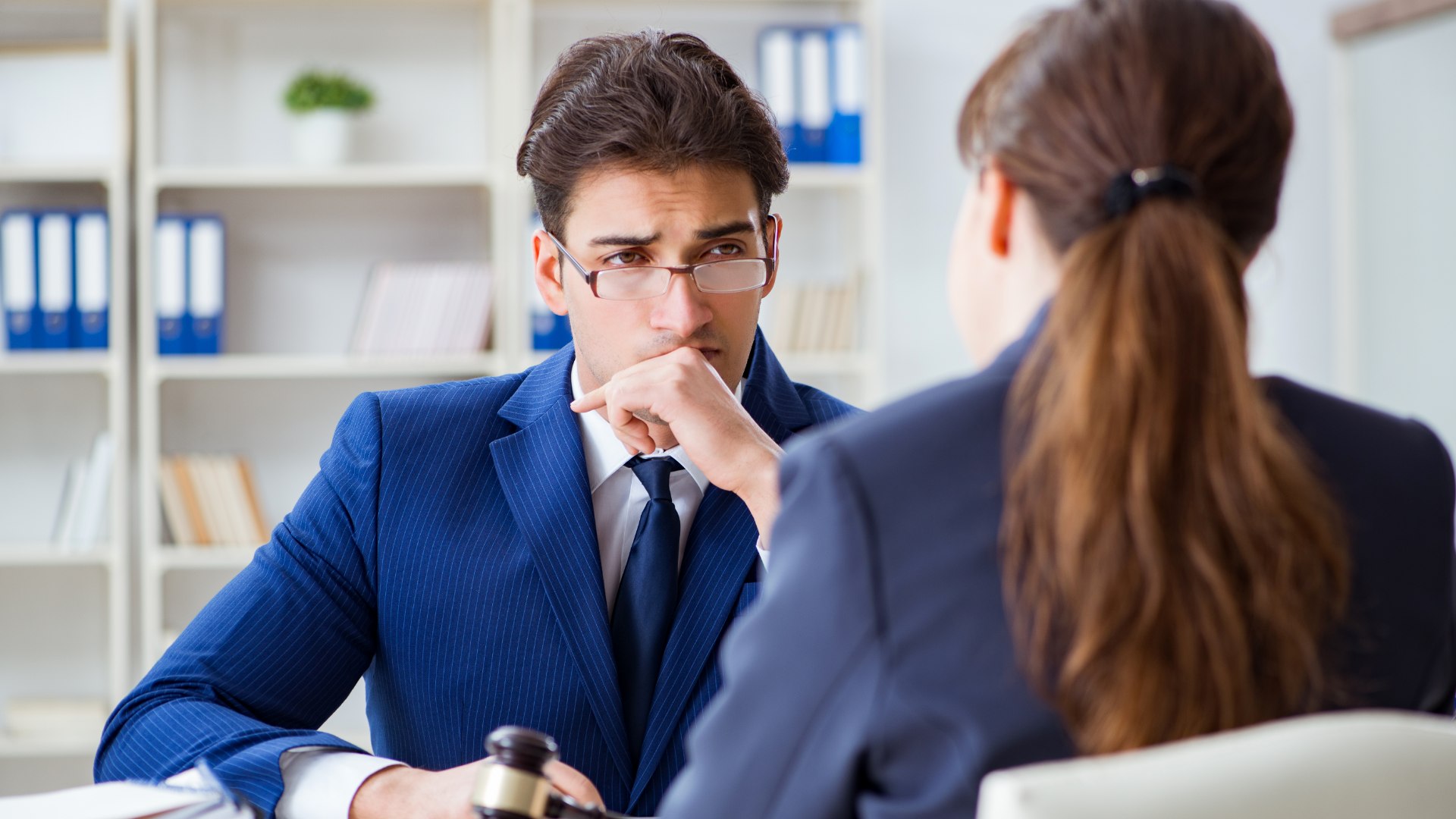 Man in blue suit, eyeglasses, looks concerned, talking to another person in a suit, in an office.