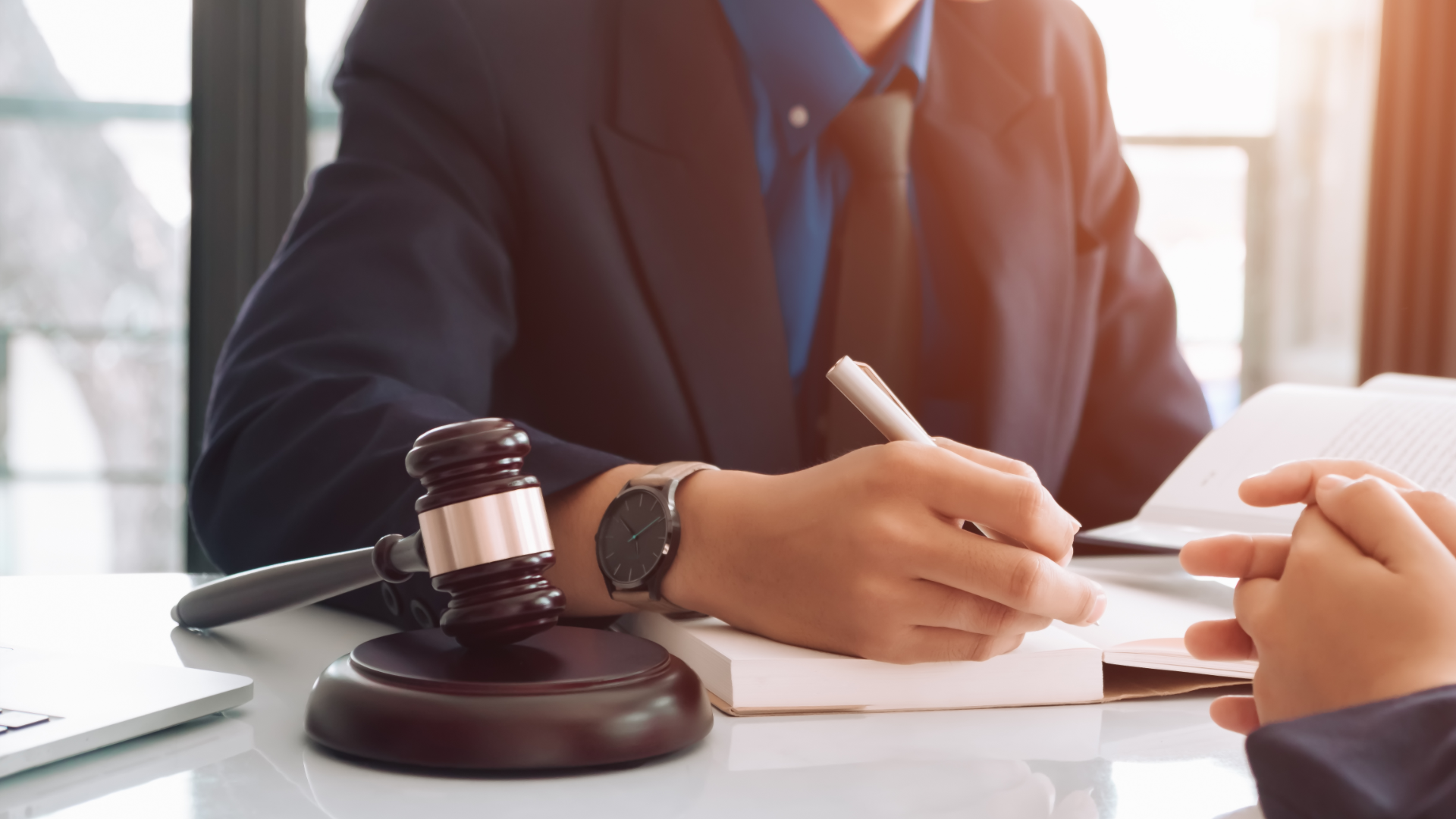 Person in suit writes in notebook at a desk with a gavel, presumably in a legal setting.