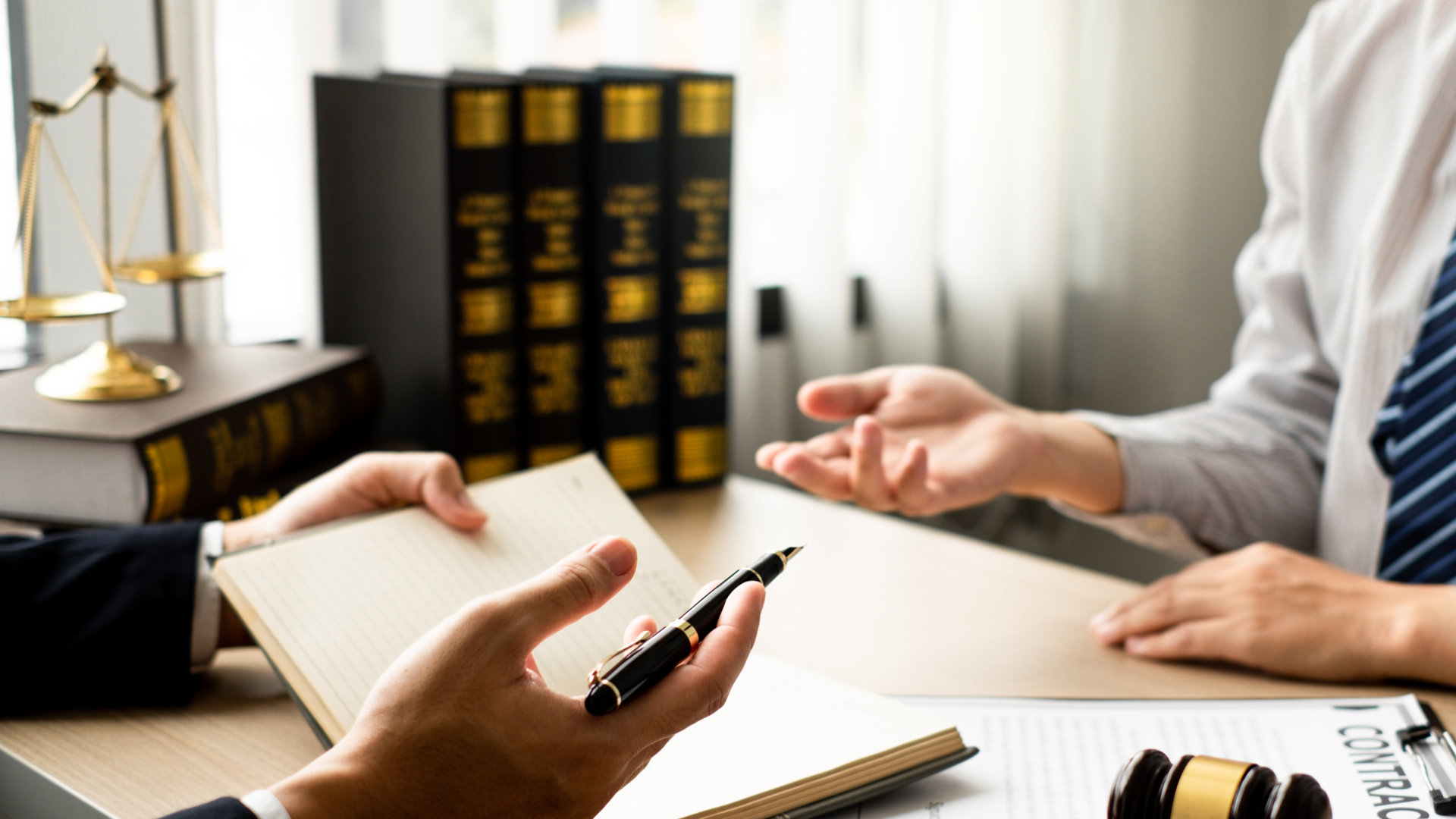 Two people in suits at a desk, reviewing documents with law books and scales of justice in the background.