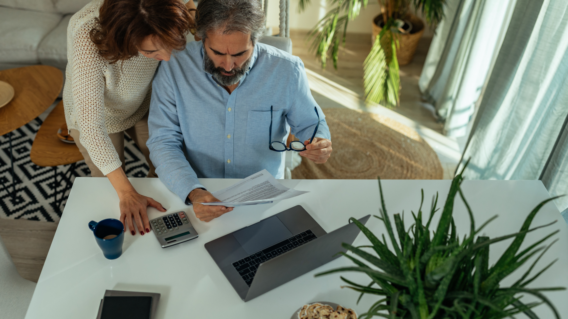 Couple reviewing documents at a table with a laptop, calculator, and plants indoors.