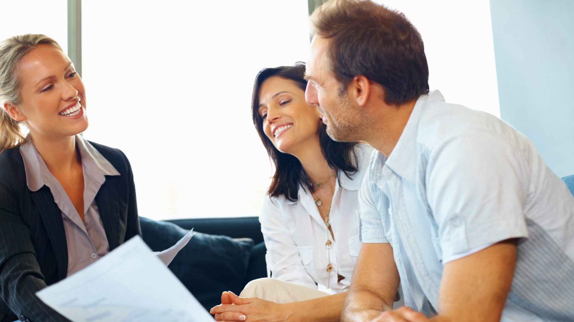Woman in suit smiles while showing papers to a smiling couple in a light-filled office.