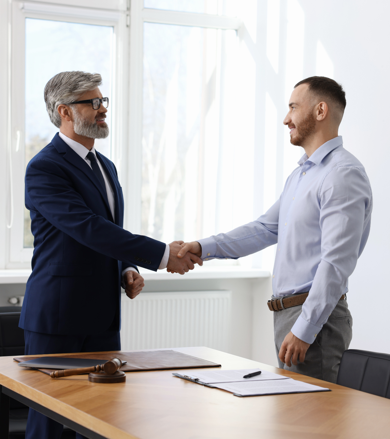 Two men shaking hands in an office, smiling. One in a suit, the other in a button-down shirt.