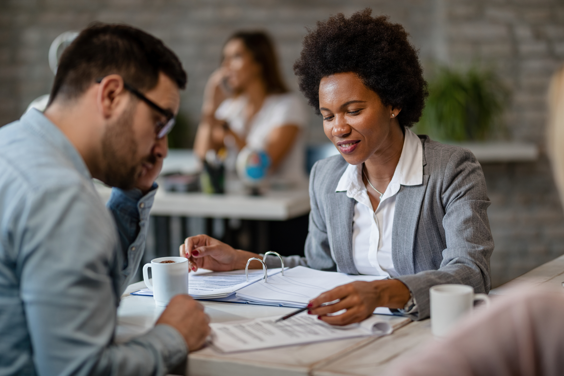 Woman in gray jacket reviews documents with a man at a table, two others in the background.