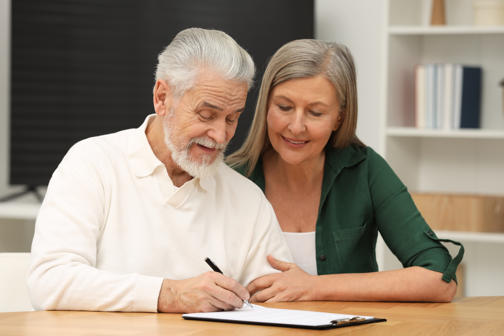 An older man signs a document at a table, watched by a woman.