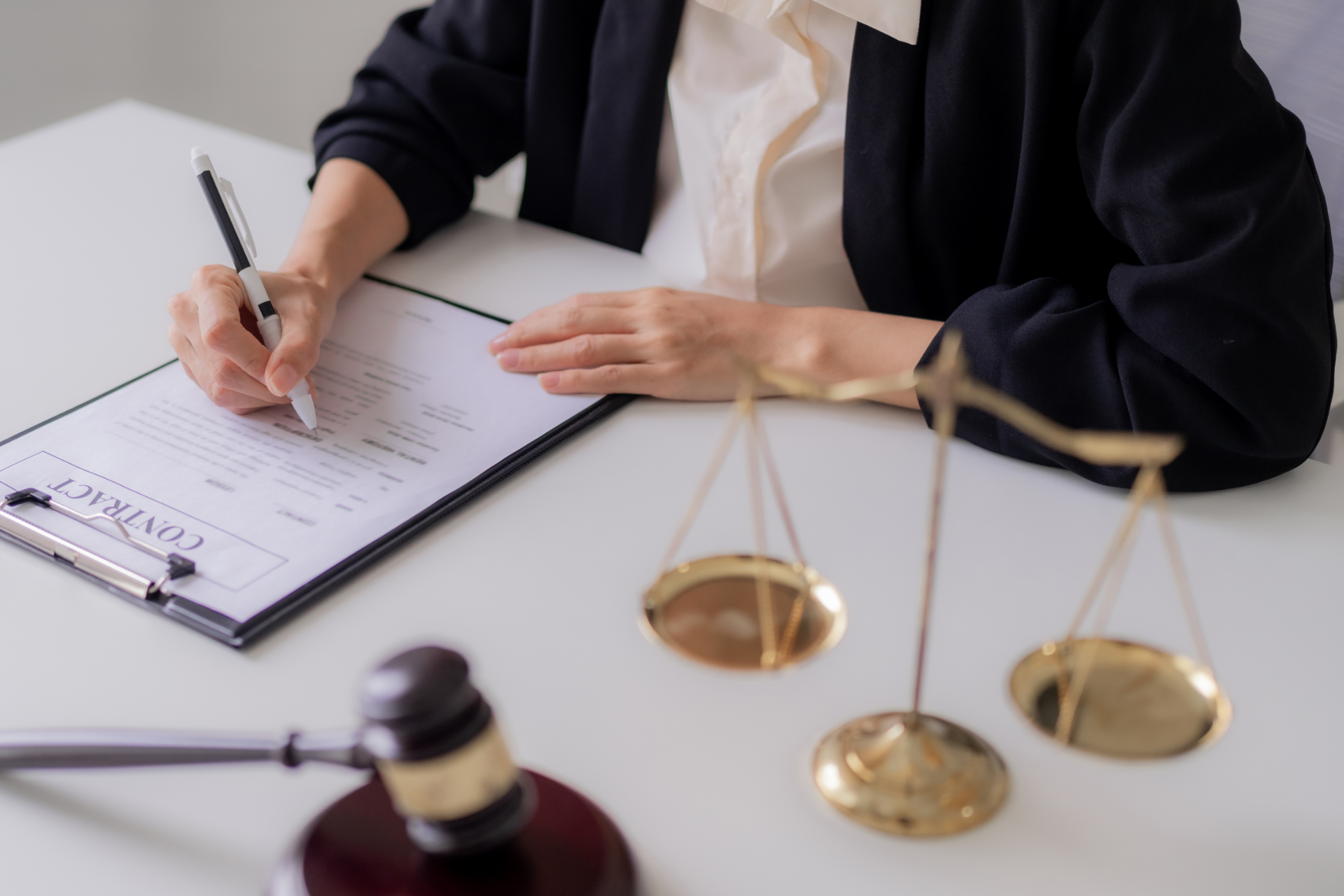 Person writing on a contract with gavel and scales of justice on a white desk.