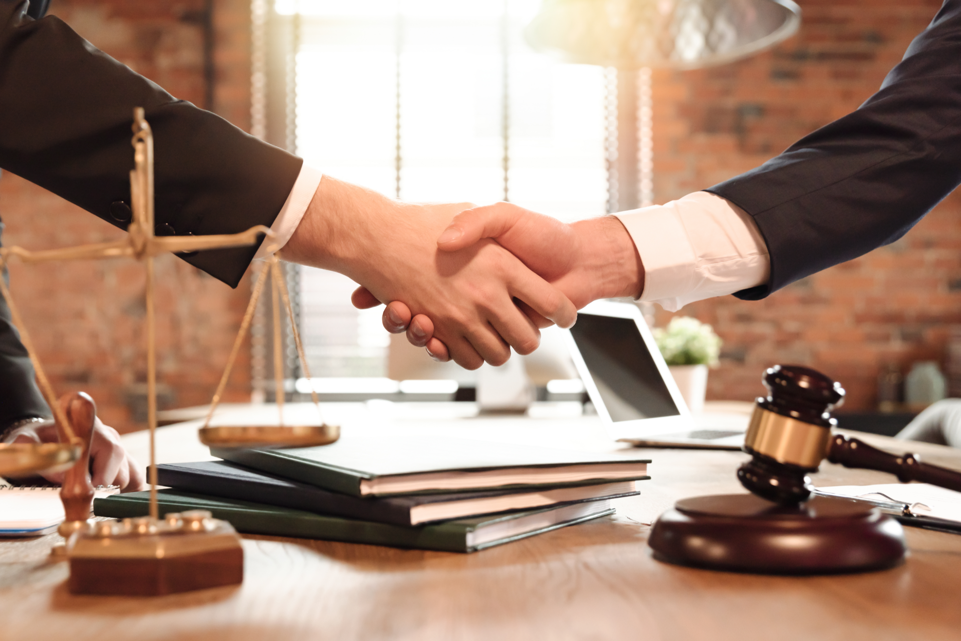 Two people in suits shaking hands over a table with a gavel, scales of justice, and documents.