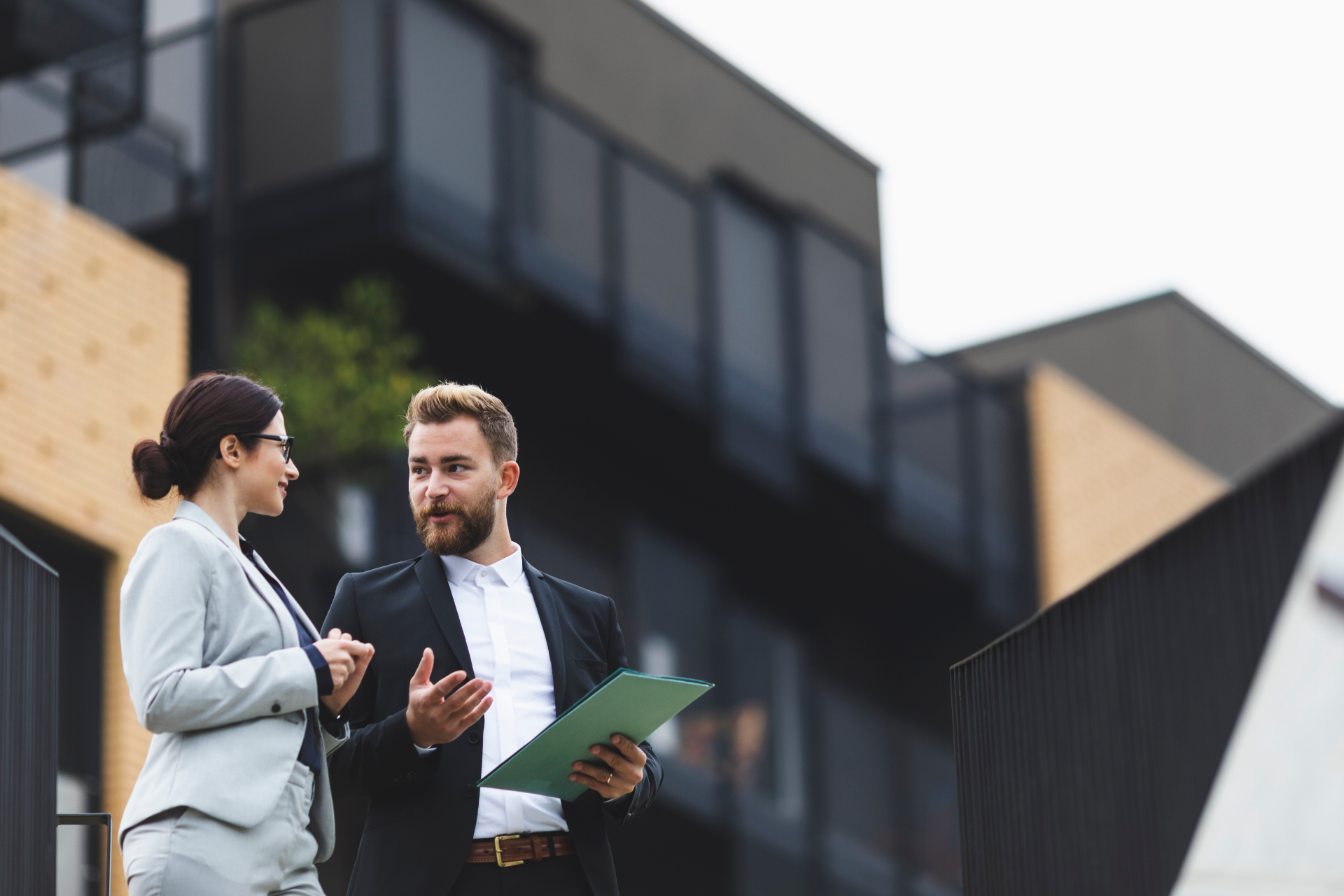 Man and woman in business attire discussing documents, modern building in background.