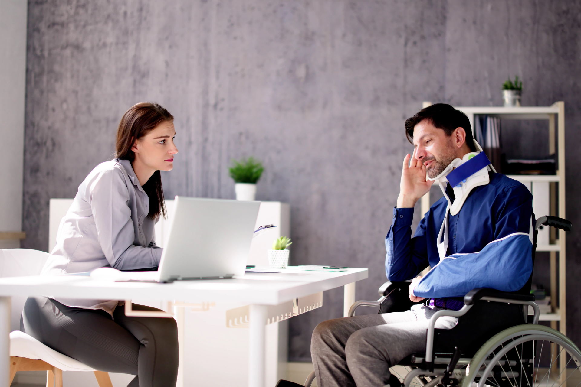 Woman with laptop talking to injured person in wheelchair, wearing arm sling and neck brace.