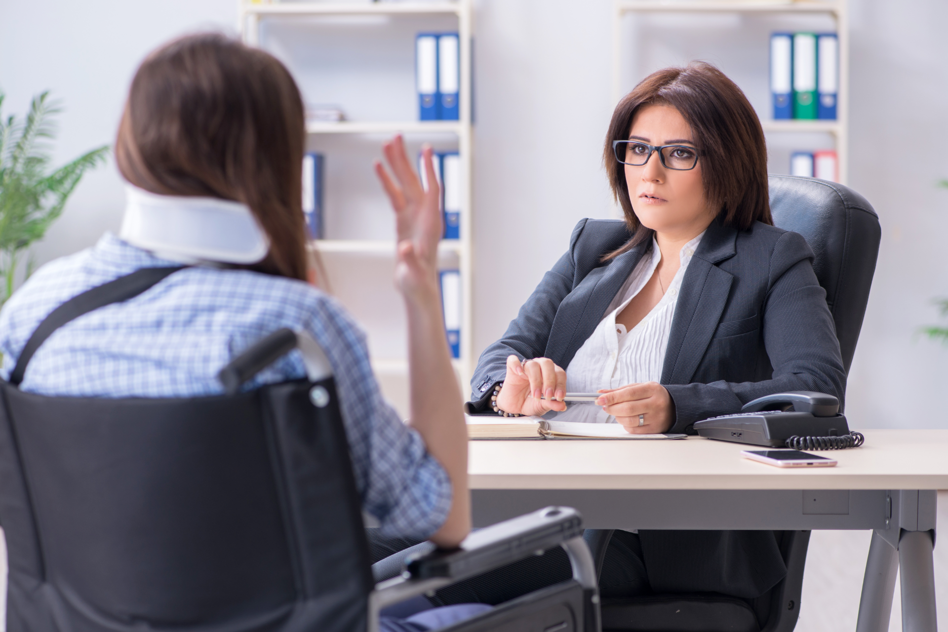 Woman in wheelchair with neck brace speaking to woman in suit at a desk. Office setting.