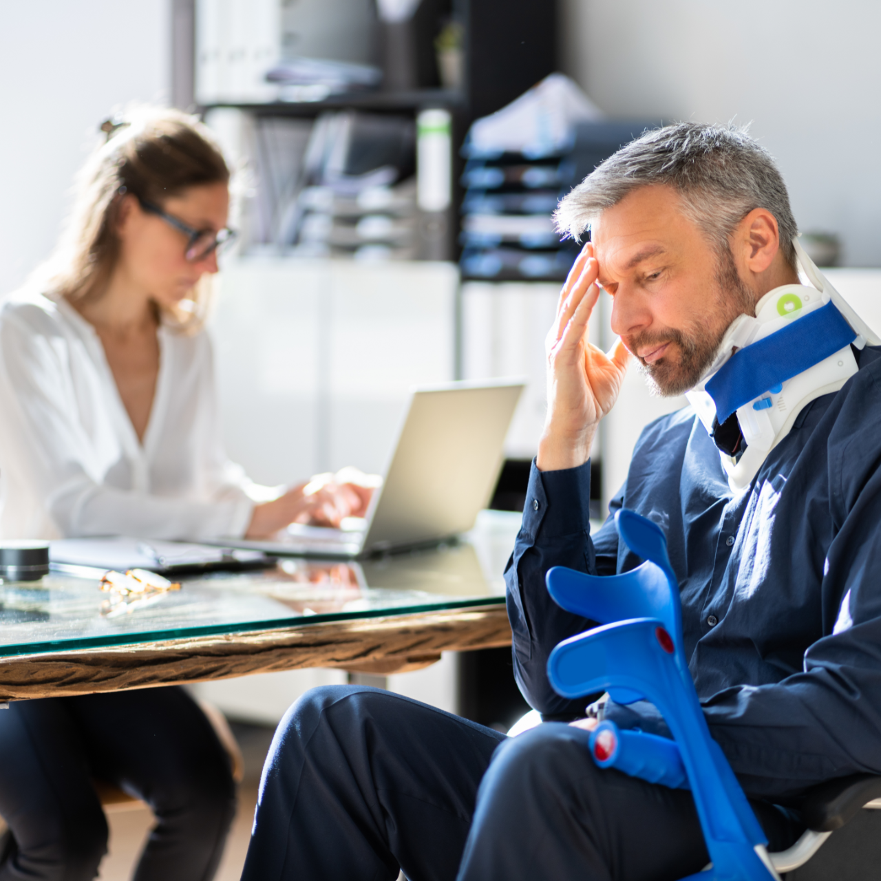 Man with neck brace and crutches at desk, looking distressed, woman working on laptop in the background.