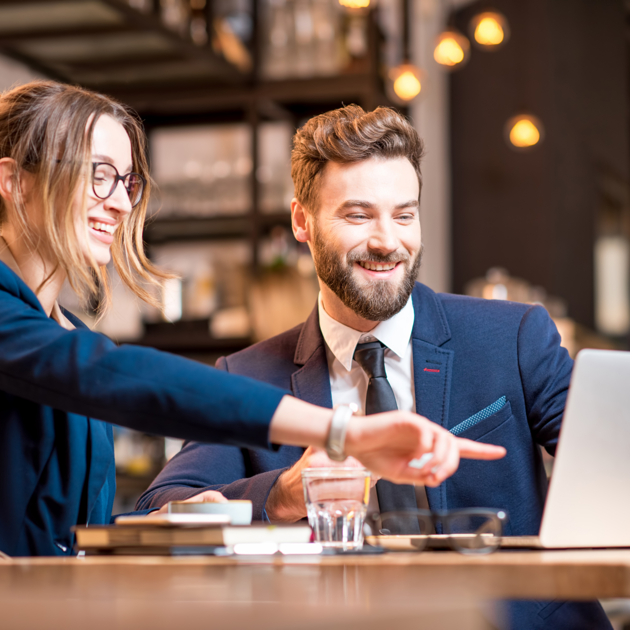 Woman points at laptop screen, smiling, while man smiles, wearing business suits. In a cafe.