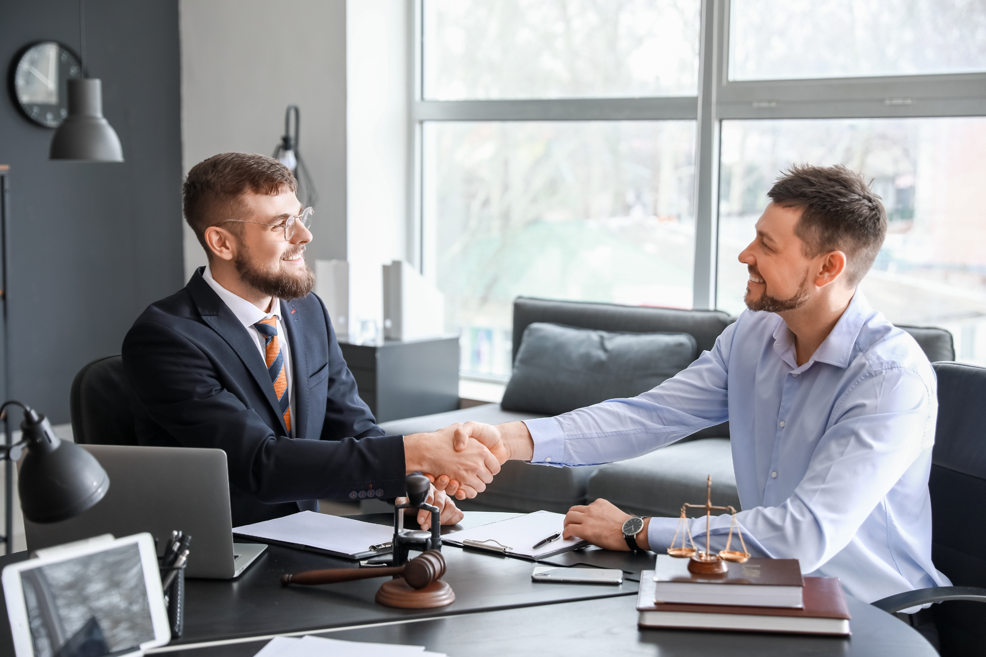 Two men shaking hands in an office, smiling. One in a suit, the other in a button-up. Desk with legal documents.
