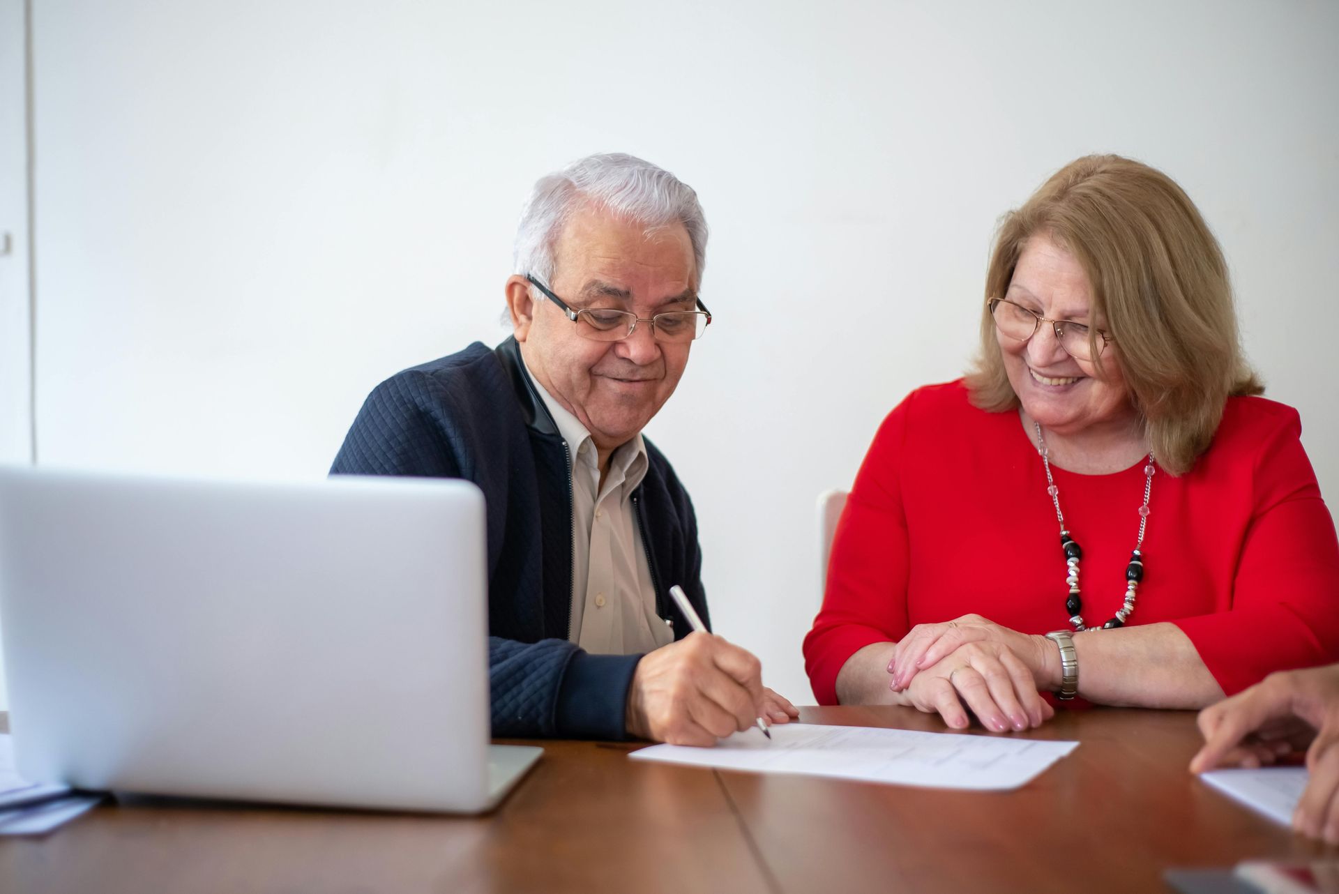 Senior couple signing documents at a table with a laptop, smiling.