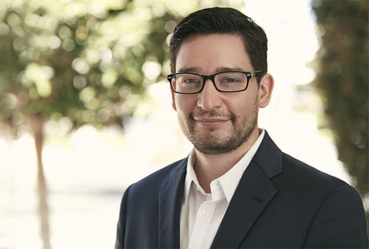 Man in glasses and suit jacket, smiling, outdoors with blurred background.