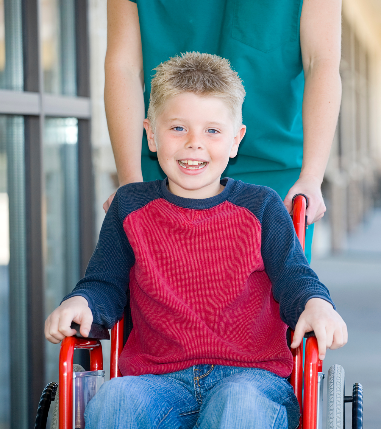 Boy in a wheelchair smiles, being pushed by a person in green scrubs outside a building.