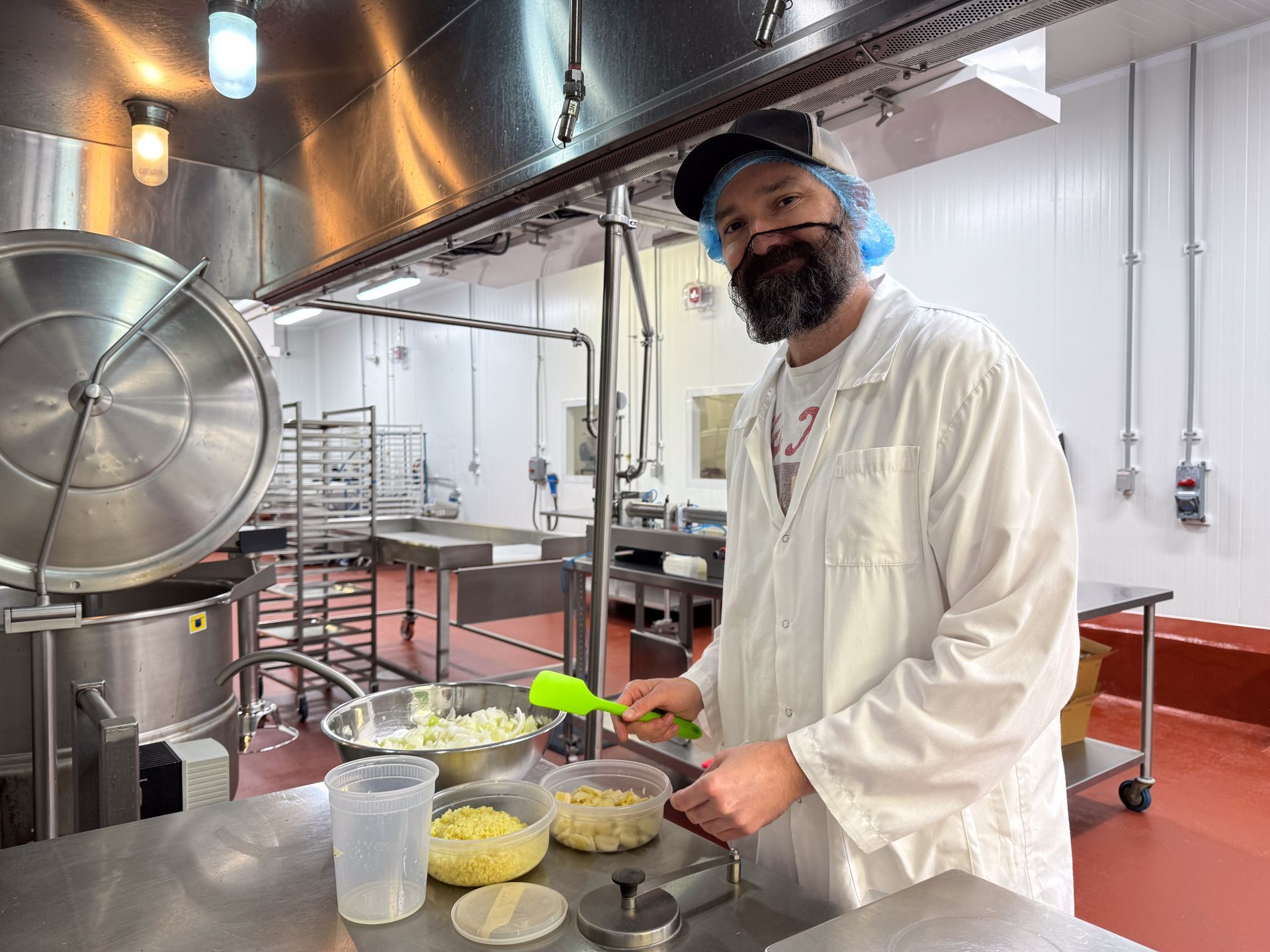 Chef in a white coat and hairnet stirring ingredients in an industrial kitchen with large metal pots and counters.
