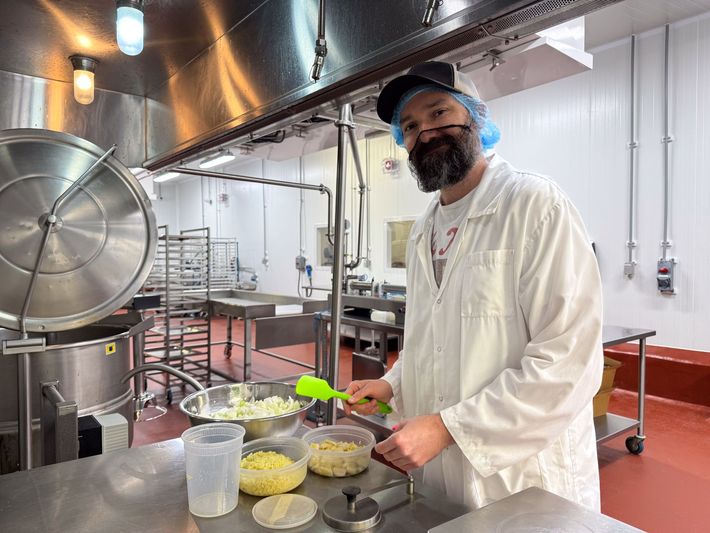Chef in a white coat and hairnet stirring ingredients in an industrial kitchen with large metal pots and counters.