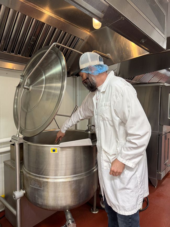 Person in white coat inspecting contents of large industrial cooking pot in a commercial kitchen.