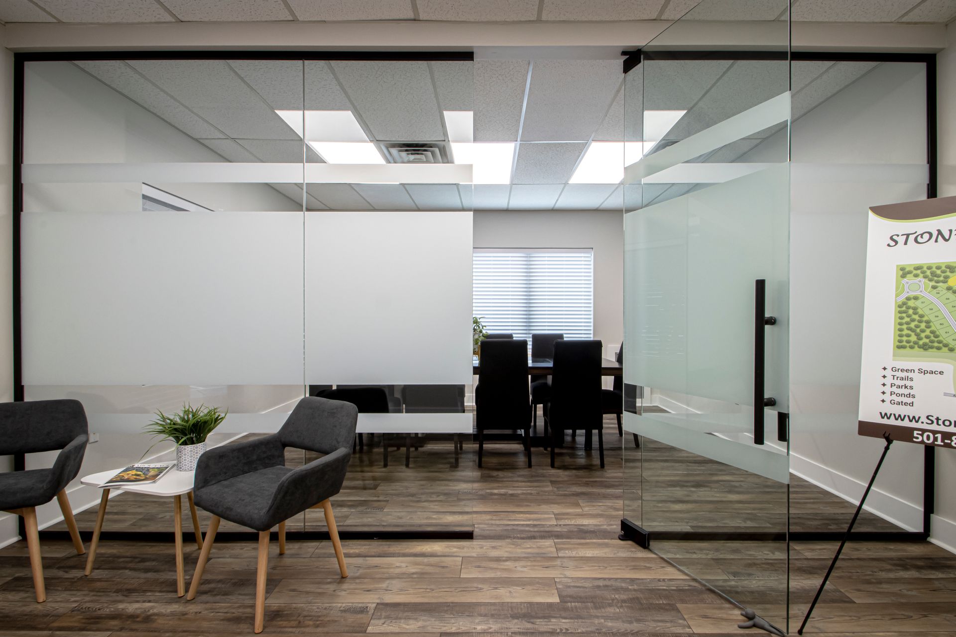 A modern office meeting room with glass walls, black chairs, a small table, and a wooden floor.