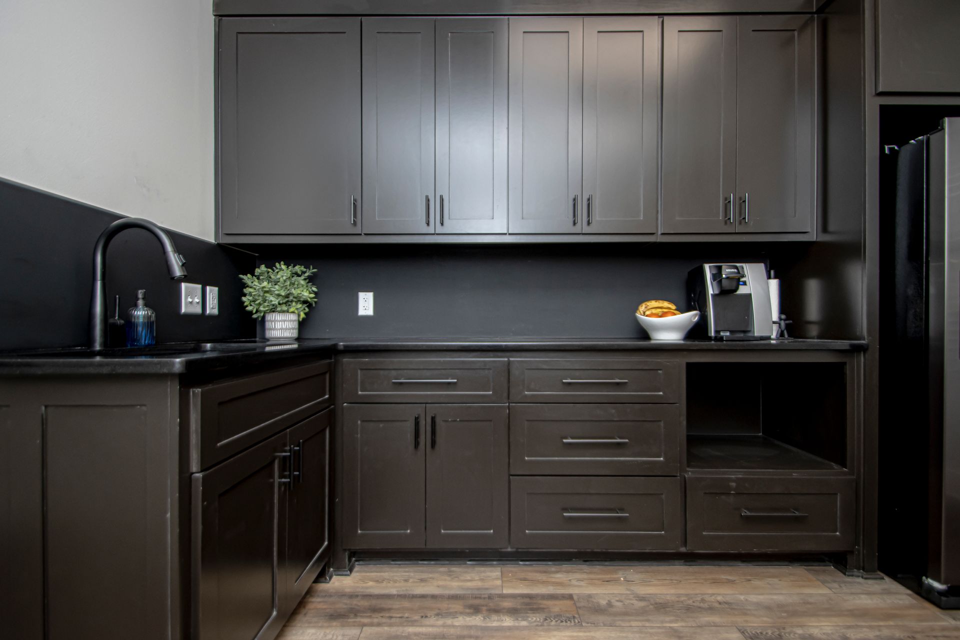 A modern kitchen wet bar featuring dark cabinetry, a black countertop, a sink, and a coffee maker against a dark wall.