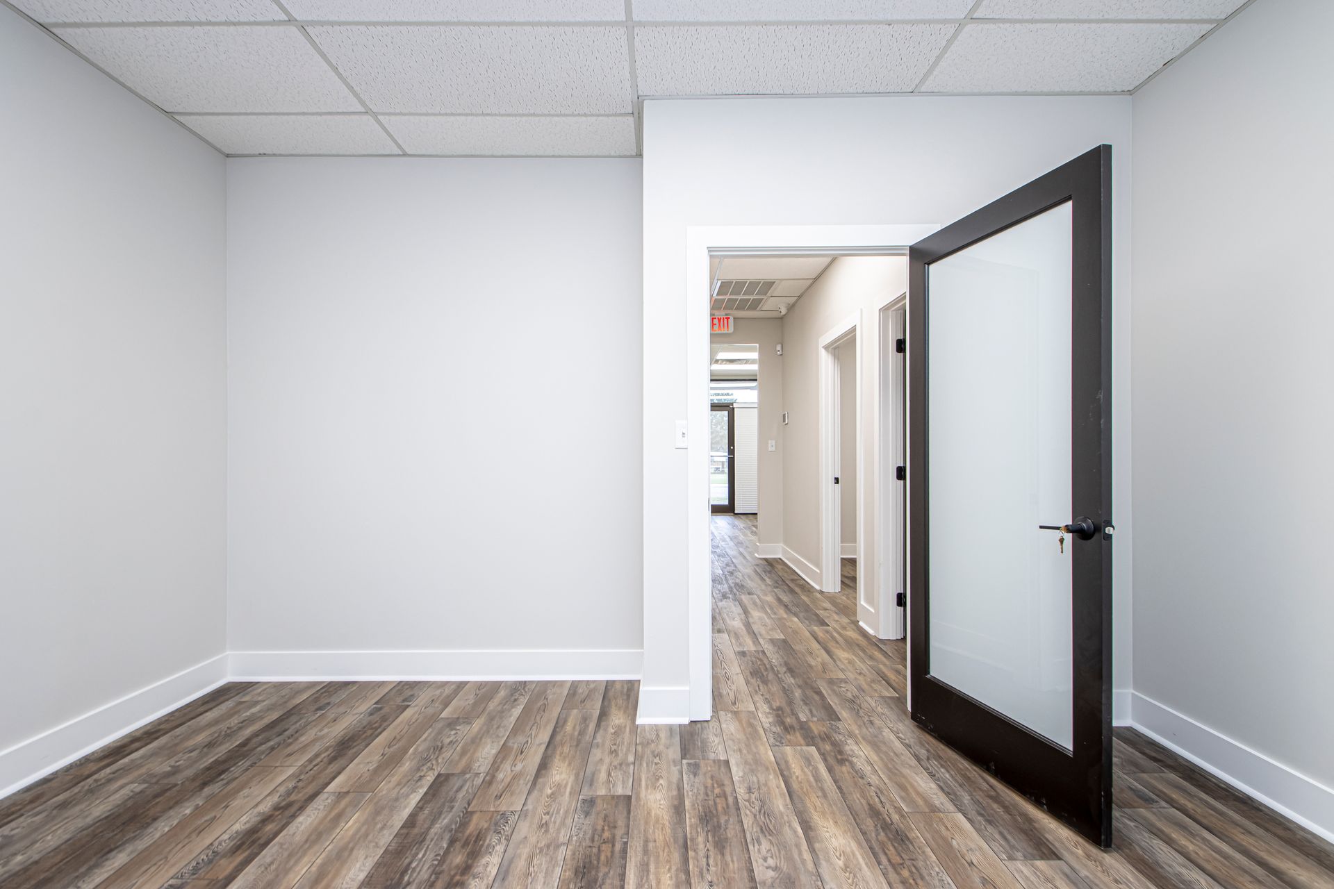 A small, empty office with light gray walls, wood-look flooring, a drop ceiling, and a glass-paneled door.