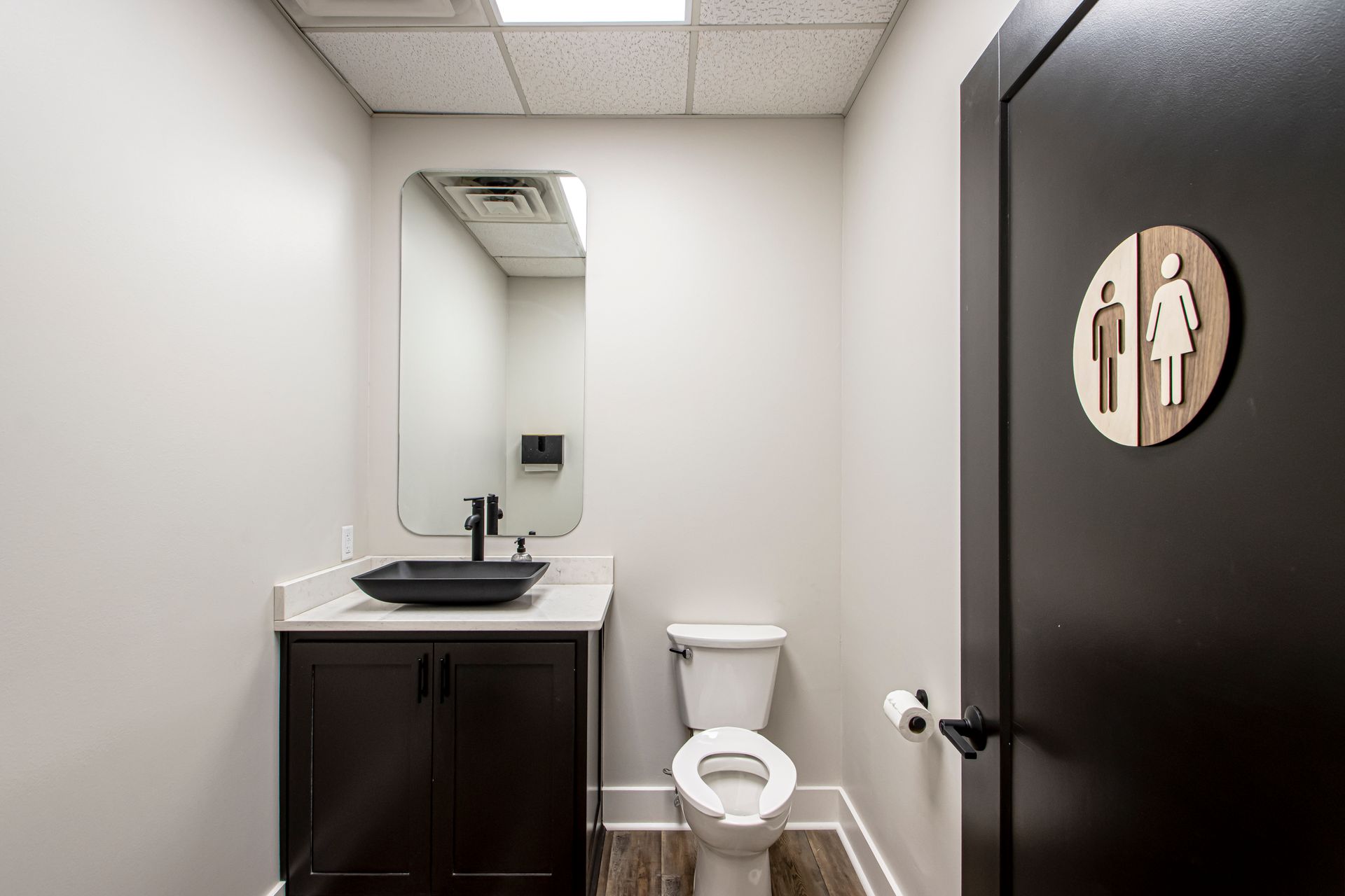 A small, neutral-toned restroom with a dark wood vanity, a black sink, a white toilet, and a circular sign on a dark door.