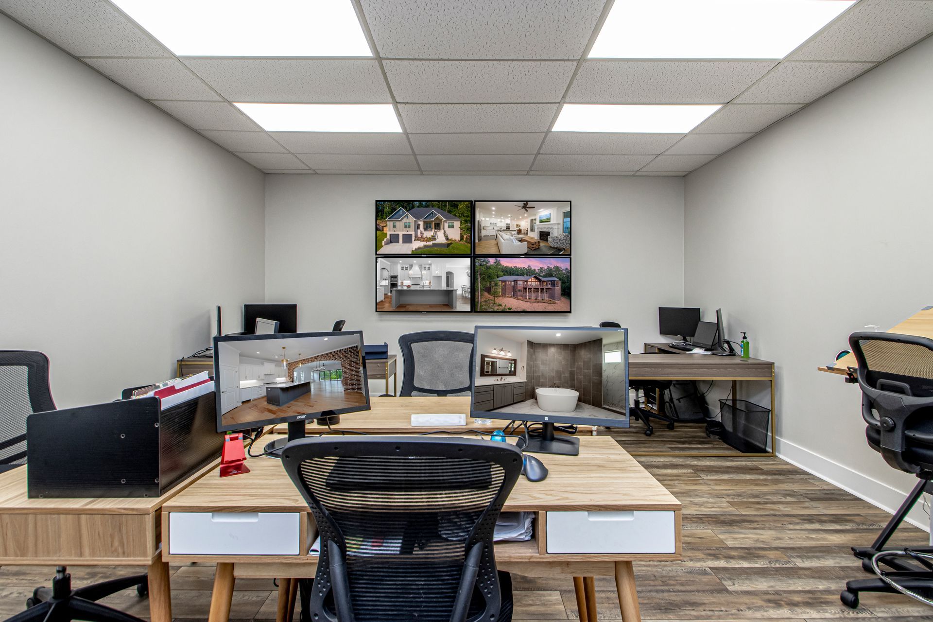 An office with two wooden desks, black chairs, and computers facing a large grid display of interior design photos.