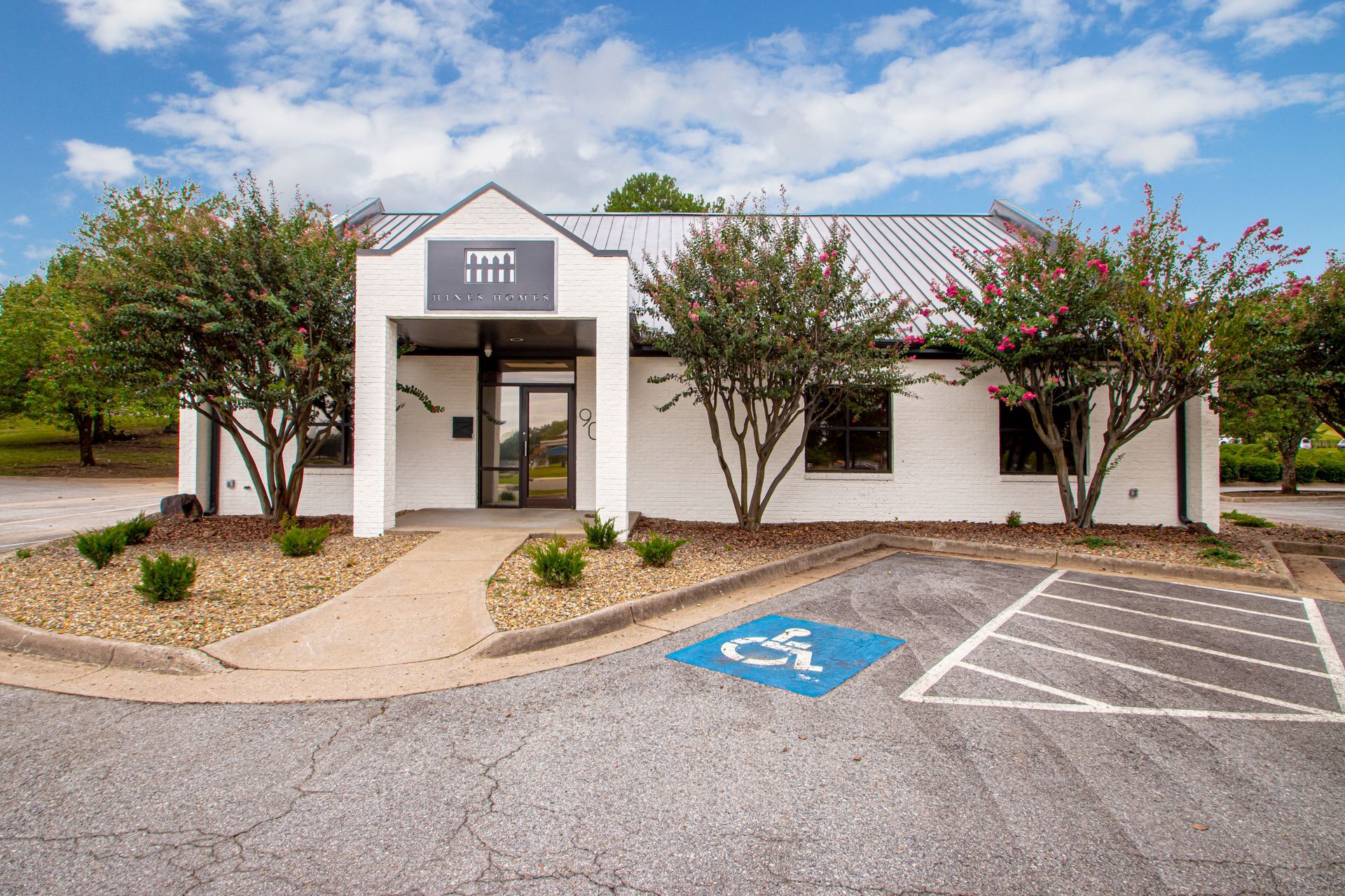 A white, single-story office building with a dark roof and trees, featuring a marked handicap parking space out front.