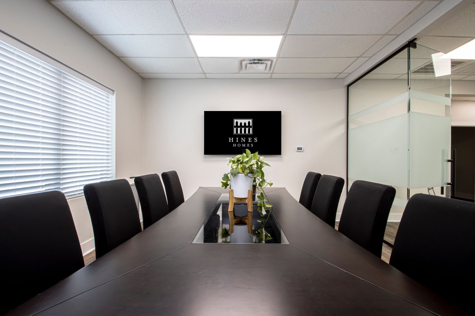 A professional conference room featuring a dark wooden table with black chairs, a centered plant, and a wall-mounted logo.