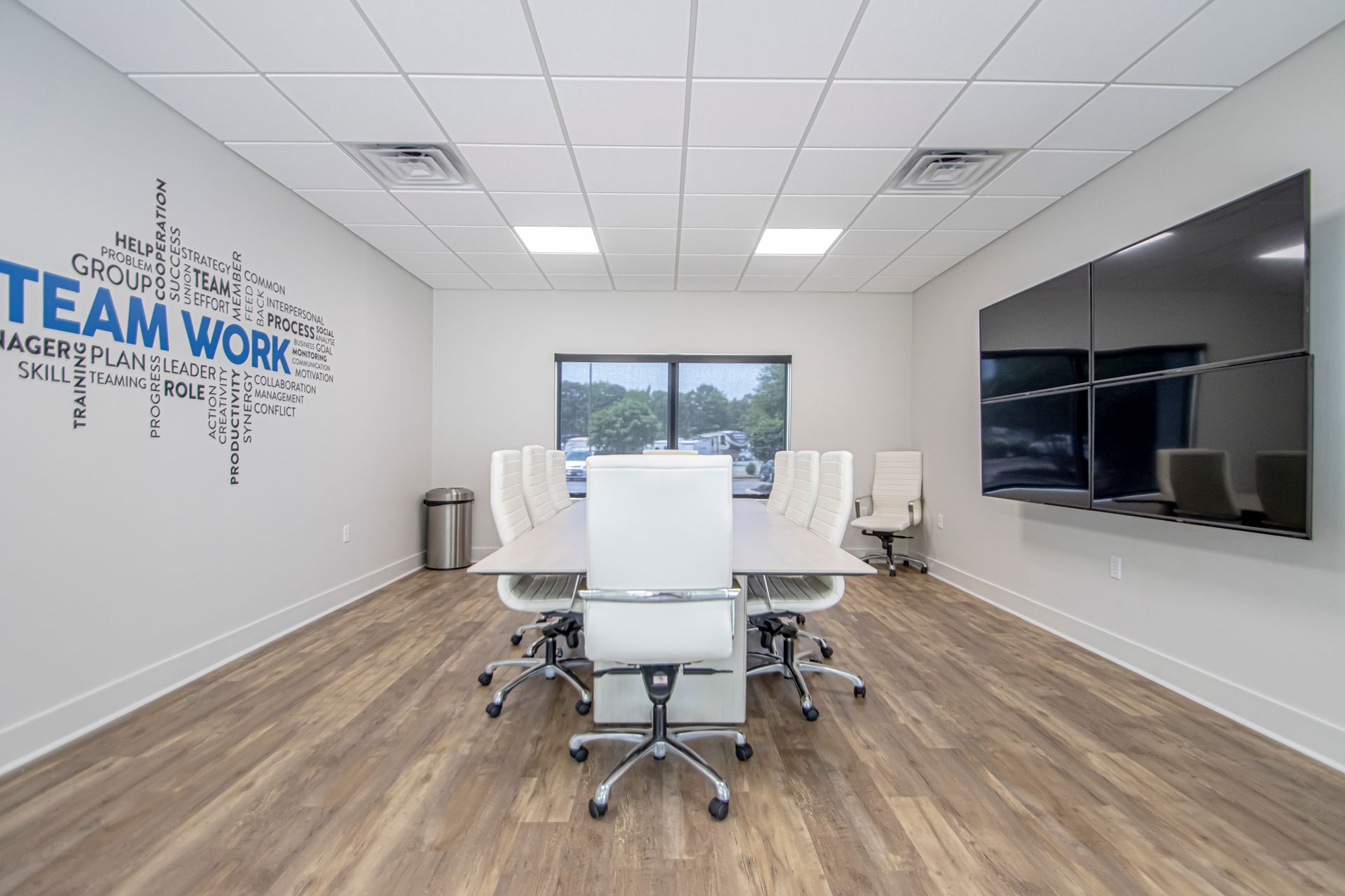 A bright office meeting room with a long table, white chairs, wood-look flooring, and a large wall-mounted monitor.
