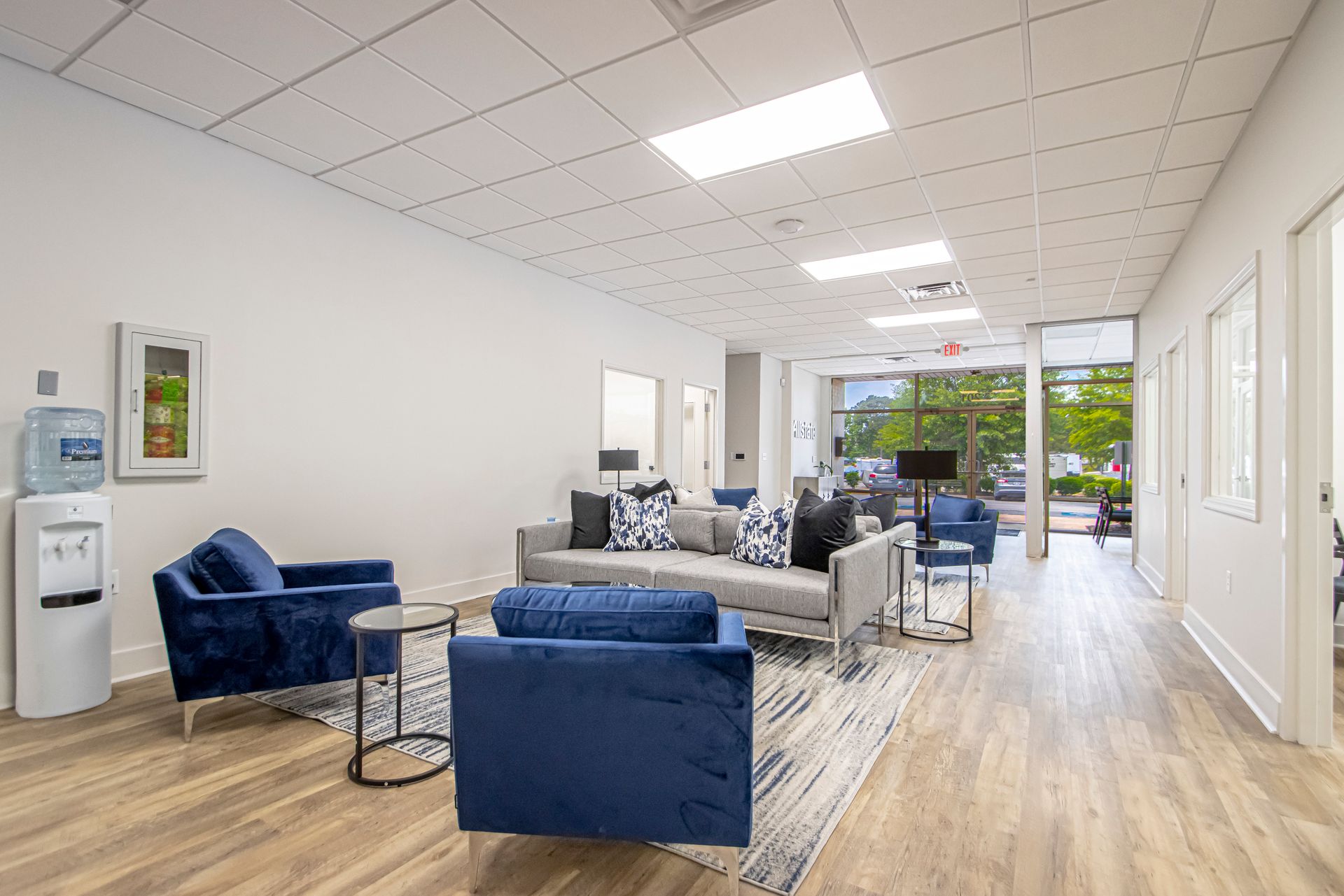 A modern, brightly lit office waiting area with blue armchairs, a gray sofa, and a water cooler on light wood flooring.