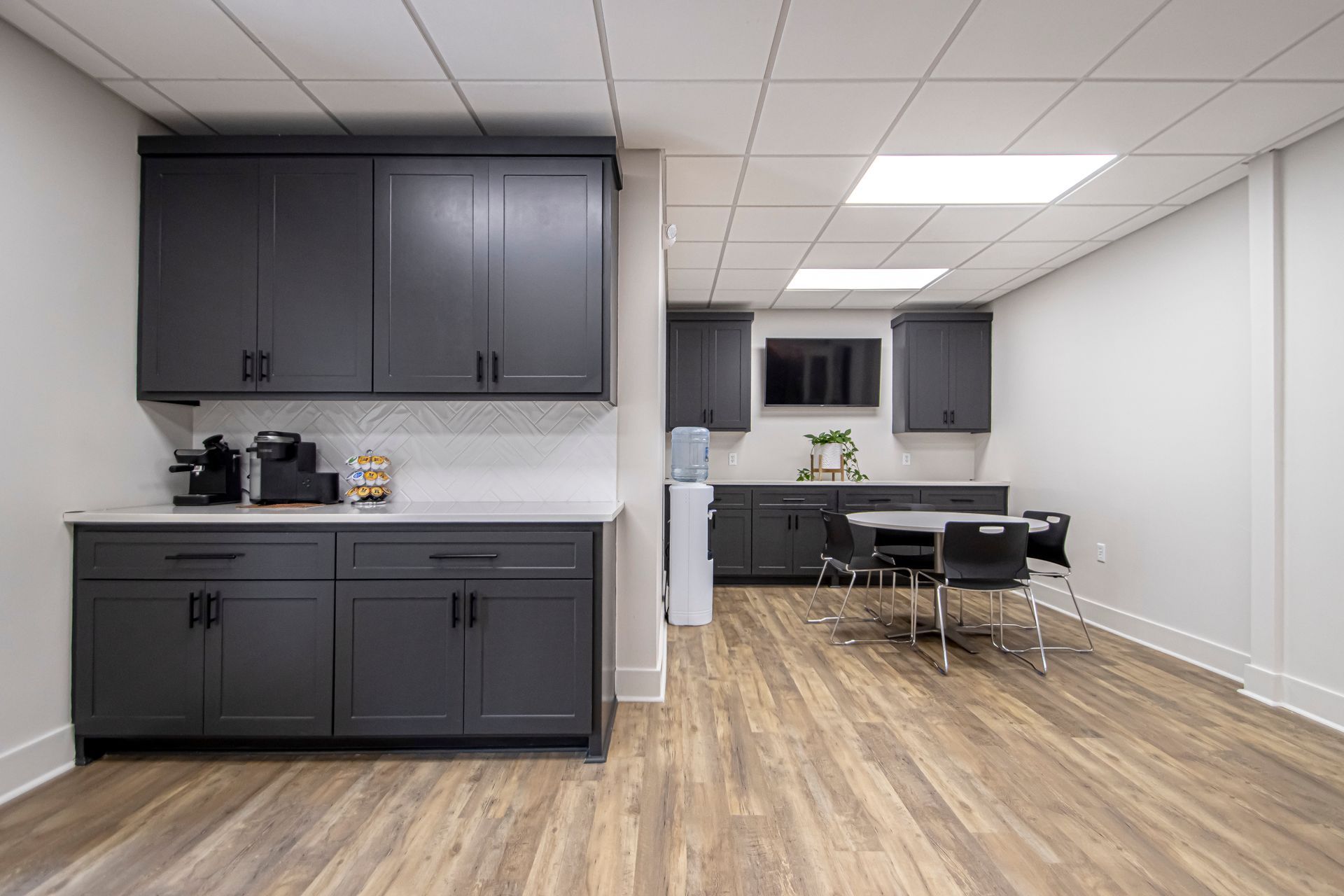A modern office breakroom with dark charcoal cabinets, white countertops, a small round table with black chairs, and TV.