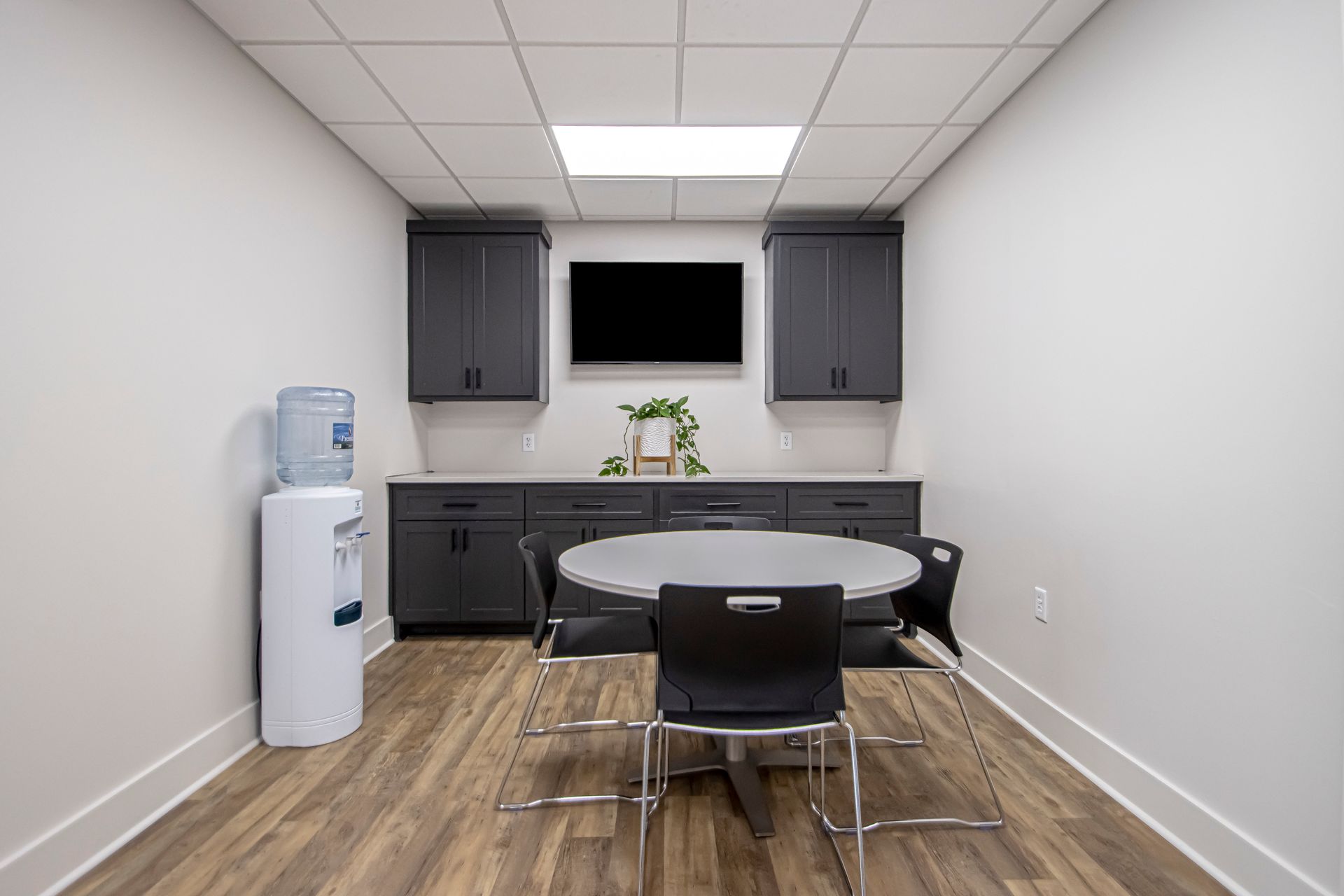 A breakroom with gray cabinets, a television, a round table with four black chairs, and a water cooler on wood floors.