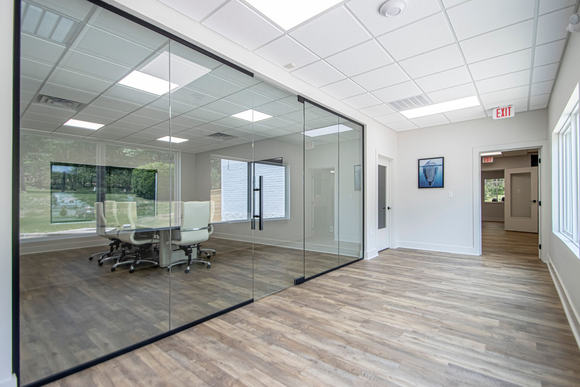 A bright office interior features a glass-walled conference room with chairs, wood flooring, and an open doorway.
