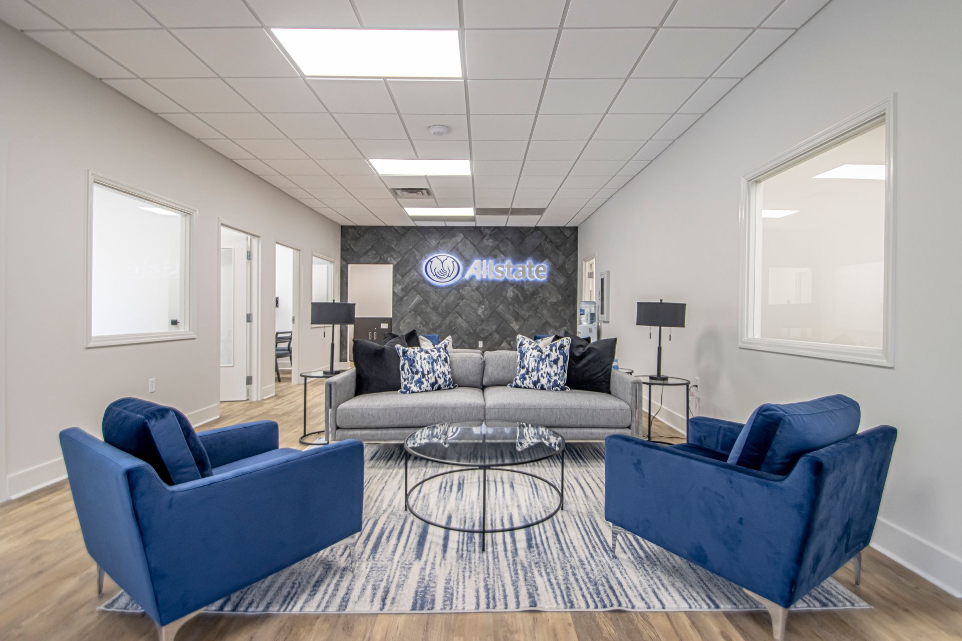 A modern office reception area featuring a grey sofa, two navy blue armchairs, and a patterned rug on a wood floor.