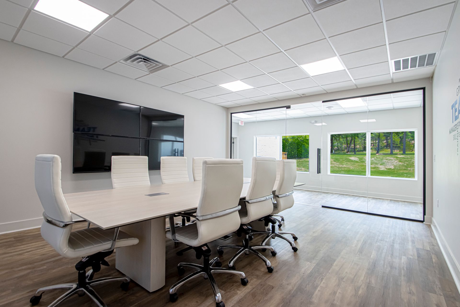 A modern, white-themed conference room with a long table, ergonomic chairs, a wall-mounted monitor, and large glass walls.