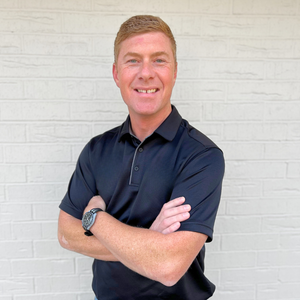 Smiling person in a navy polo shirt standing with arms crossed against a white brick wall