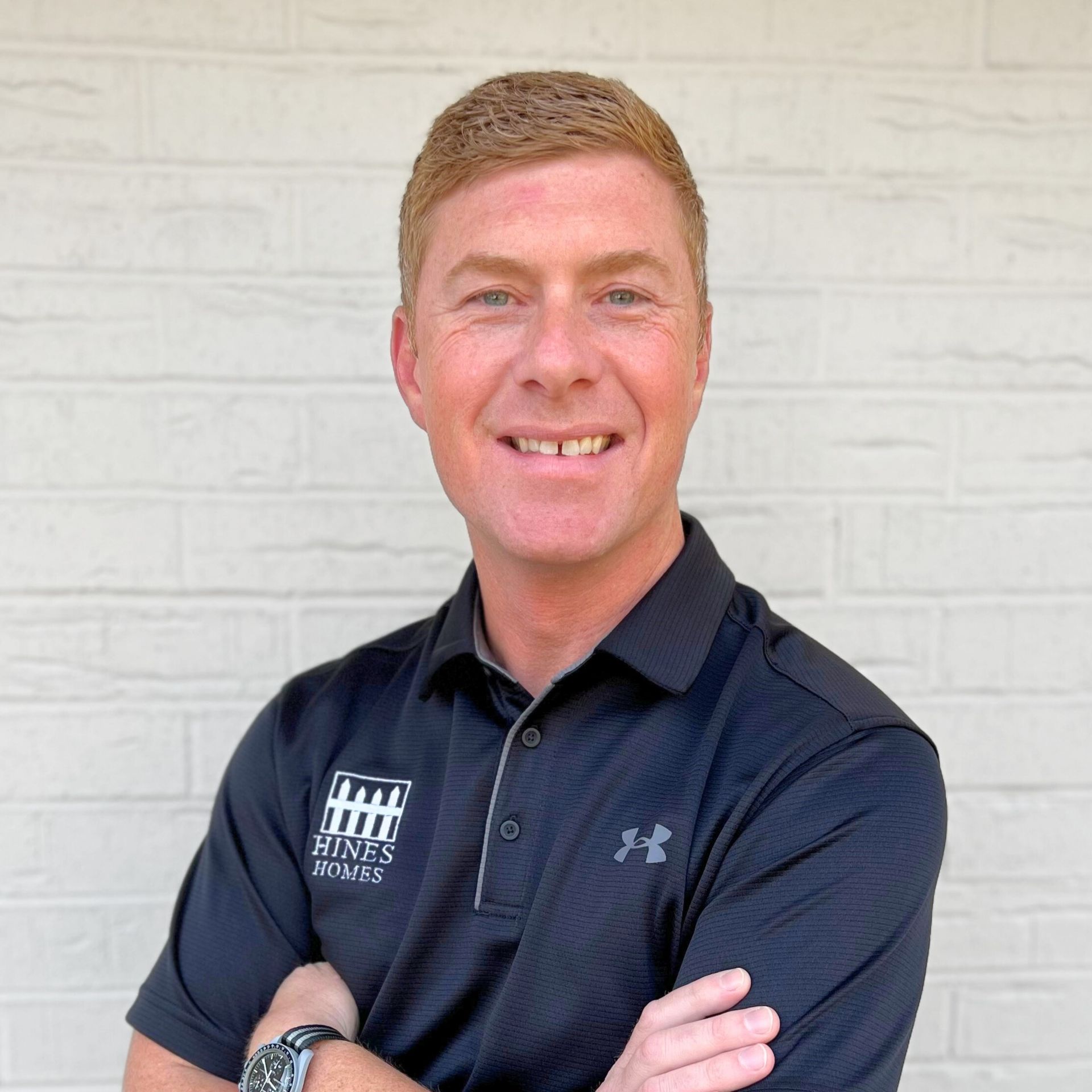 A person smiling while wearing a black polo shirt with a company logo, standing with arms crossed against a brick wall.