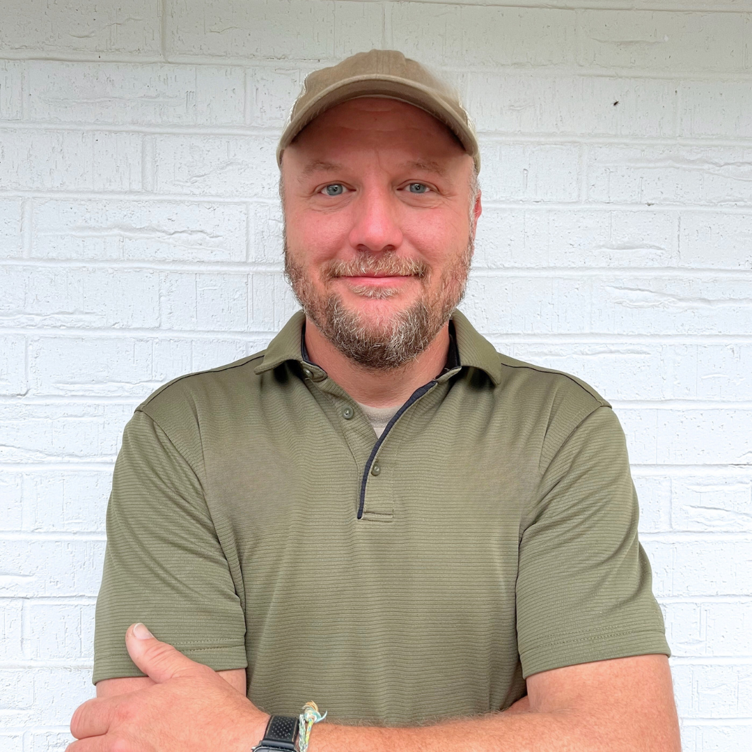 A person wearing a green polo shirt and a matching baseball cap standing against a white brick wall.
