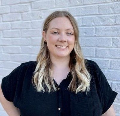 A smiling individual with shoulder-length blonde hair, wearing a black shirt, posing in front of a white brick wall.