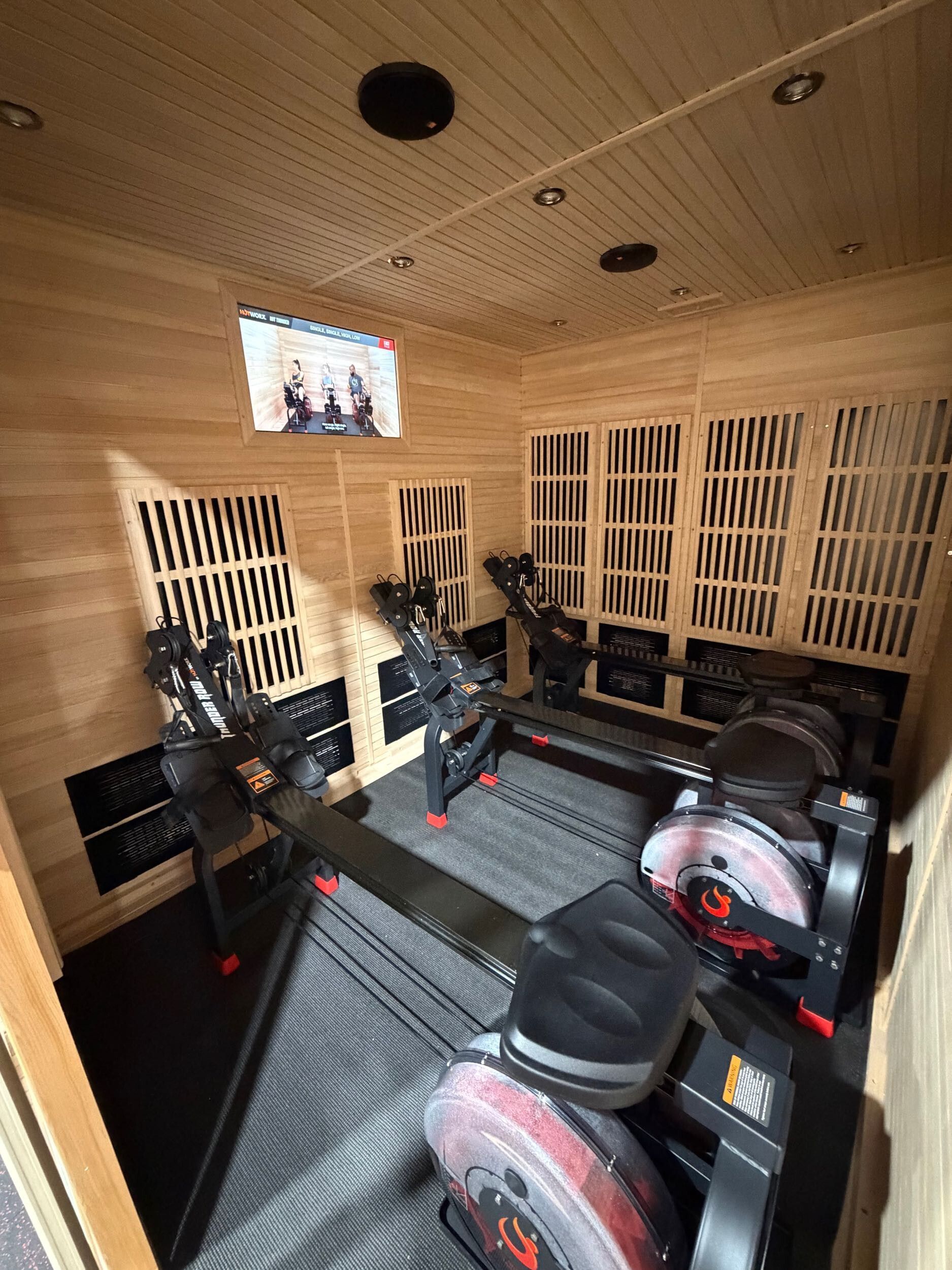 Two indoor rowing machines arranged side-by-side inside a wooden infrared sauna with a small television on the back wall.