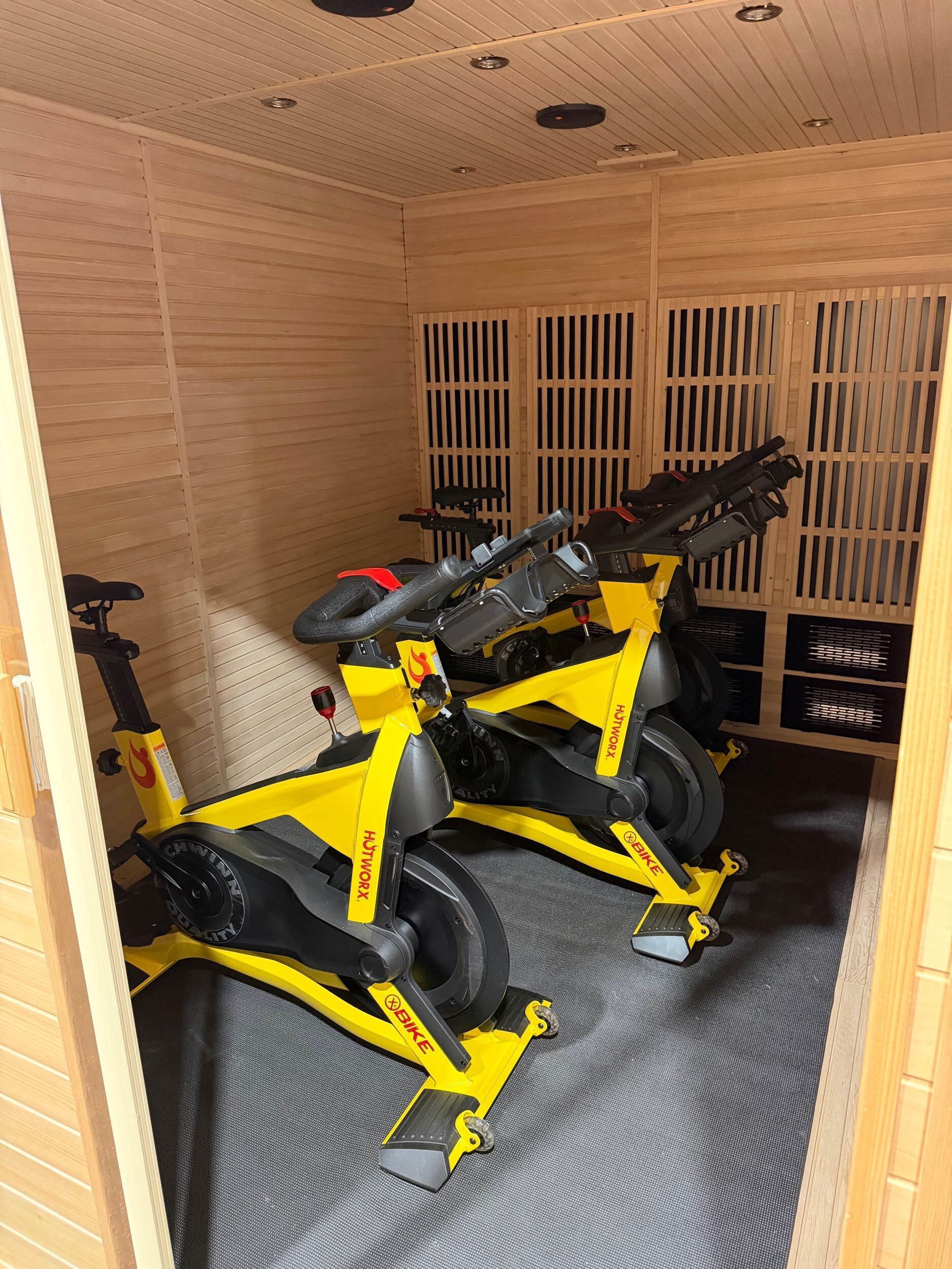 Two bright yellow stationary exercise bikes inside a wood-paneled sauna room with slatted walls.