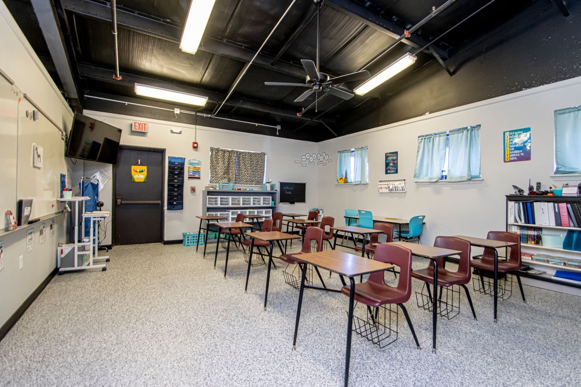 Empty classroom with rows of desks, white walls, black ceiling, tiled floor, and storage shelves.