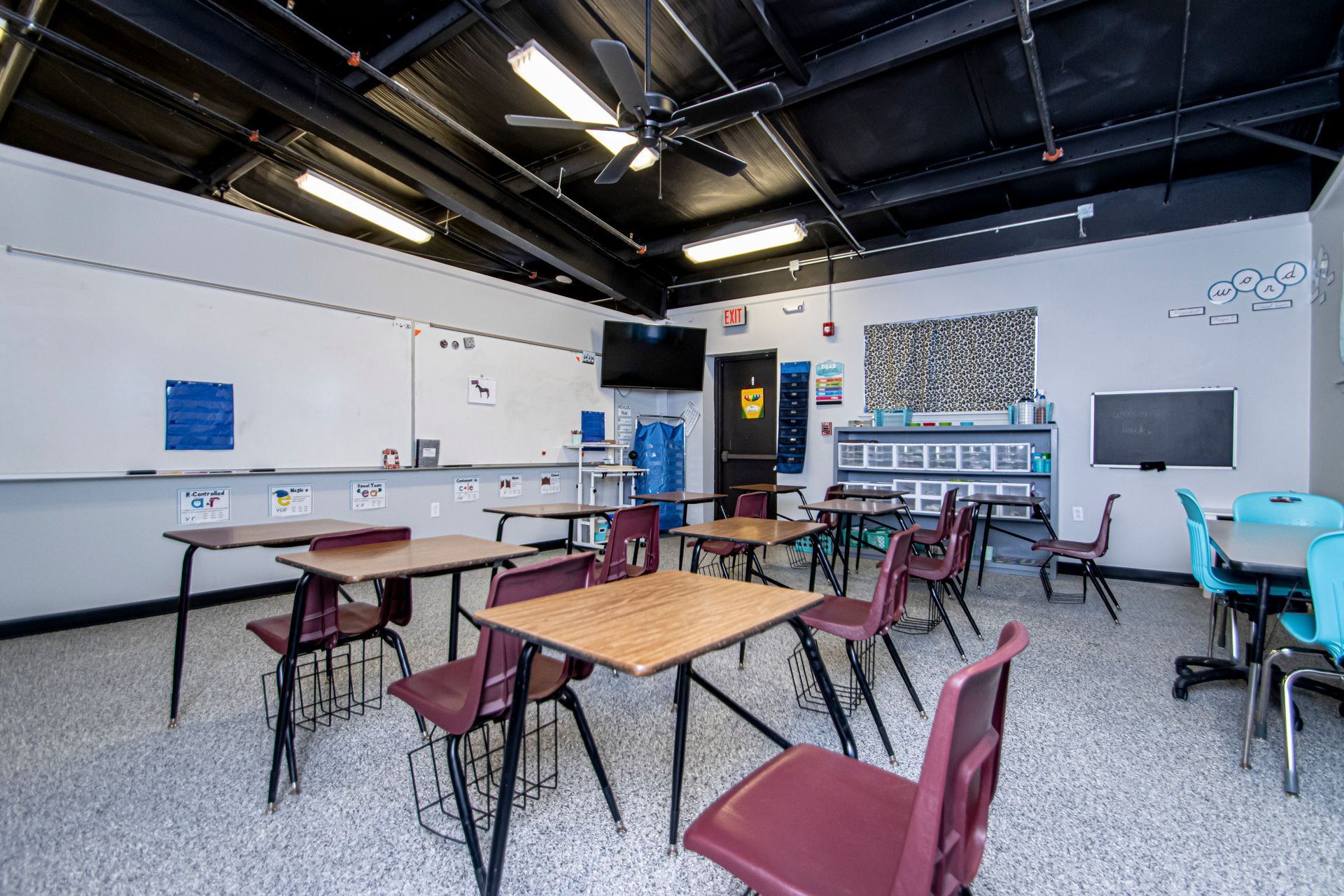 A classroom with rows of desks and maroon chairs facing whiteboards, under a black ceiling with fluorescent lighting.