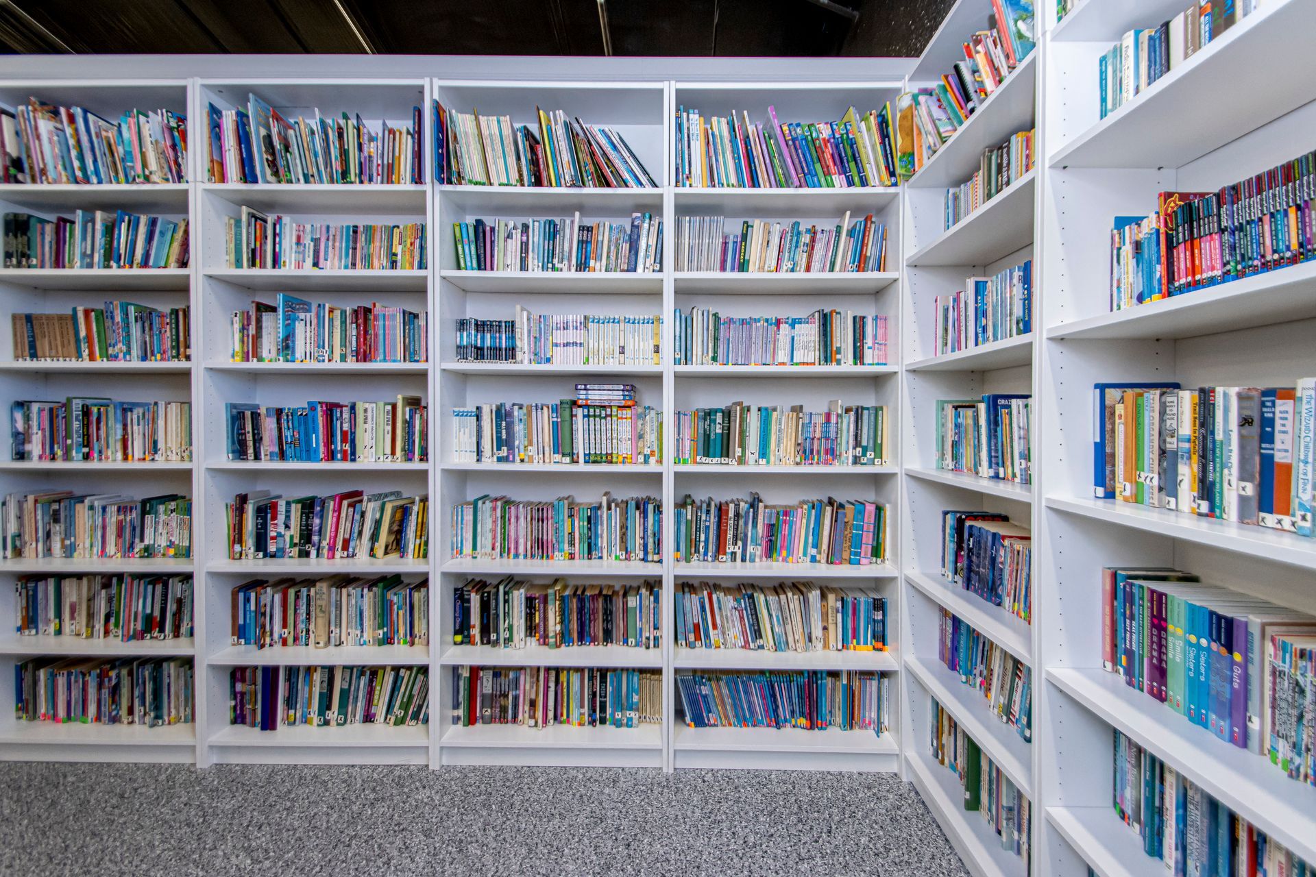 White bookshelves filled with numerous colorful books arranged on multiple rows in a brightly lit library.