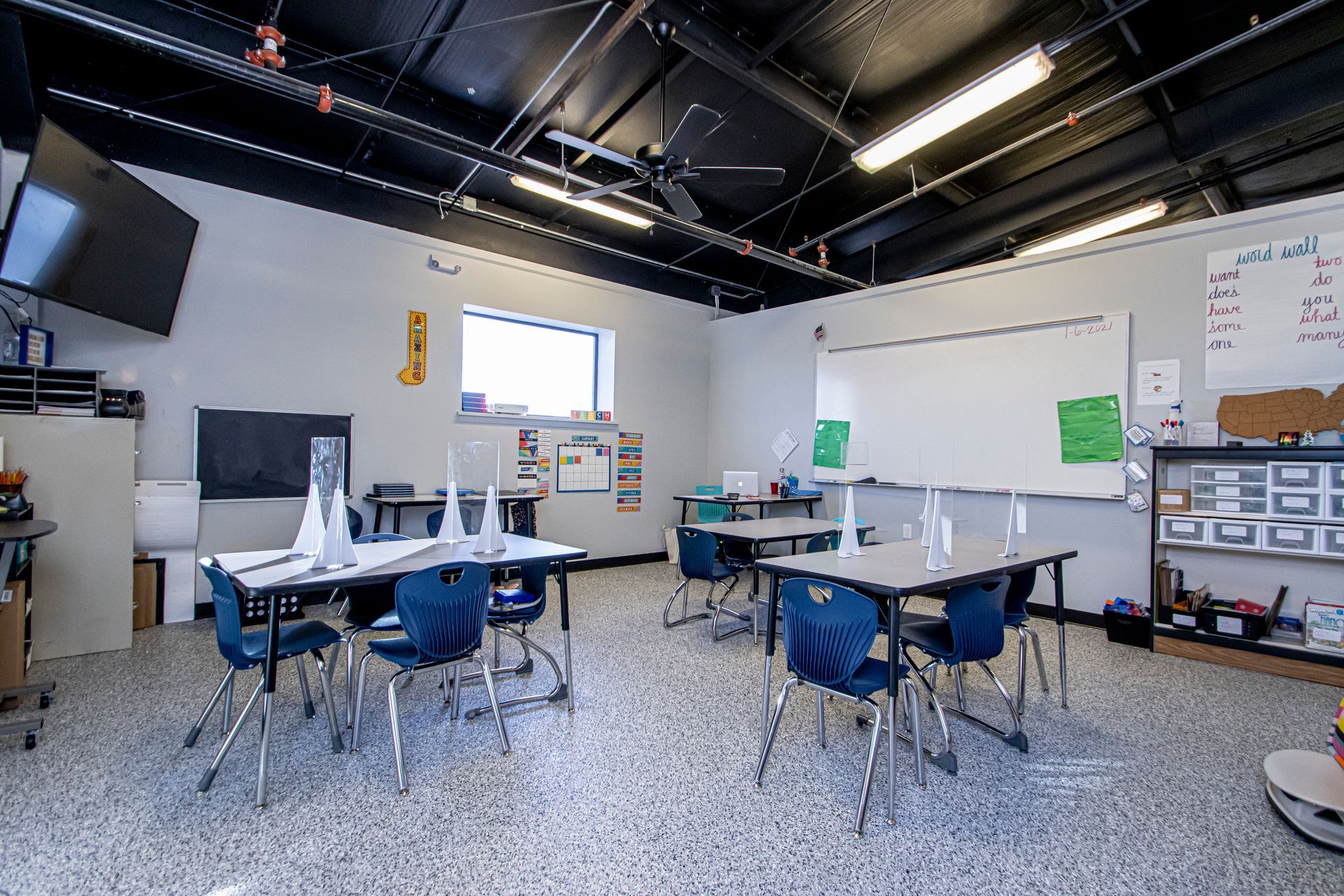 Empty classroom with tables, blue chairs, a whiteboard, and high, black-painted exposed ceilings.