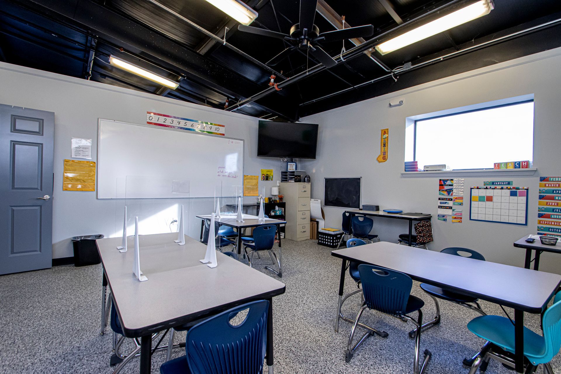 An empty classroom with desks, chairs, a whiteboard, a television, and large windows under a black industrial ceiling.