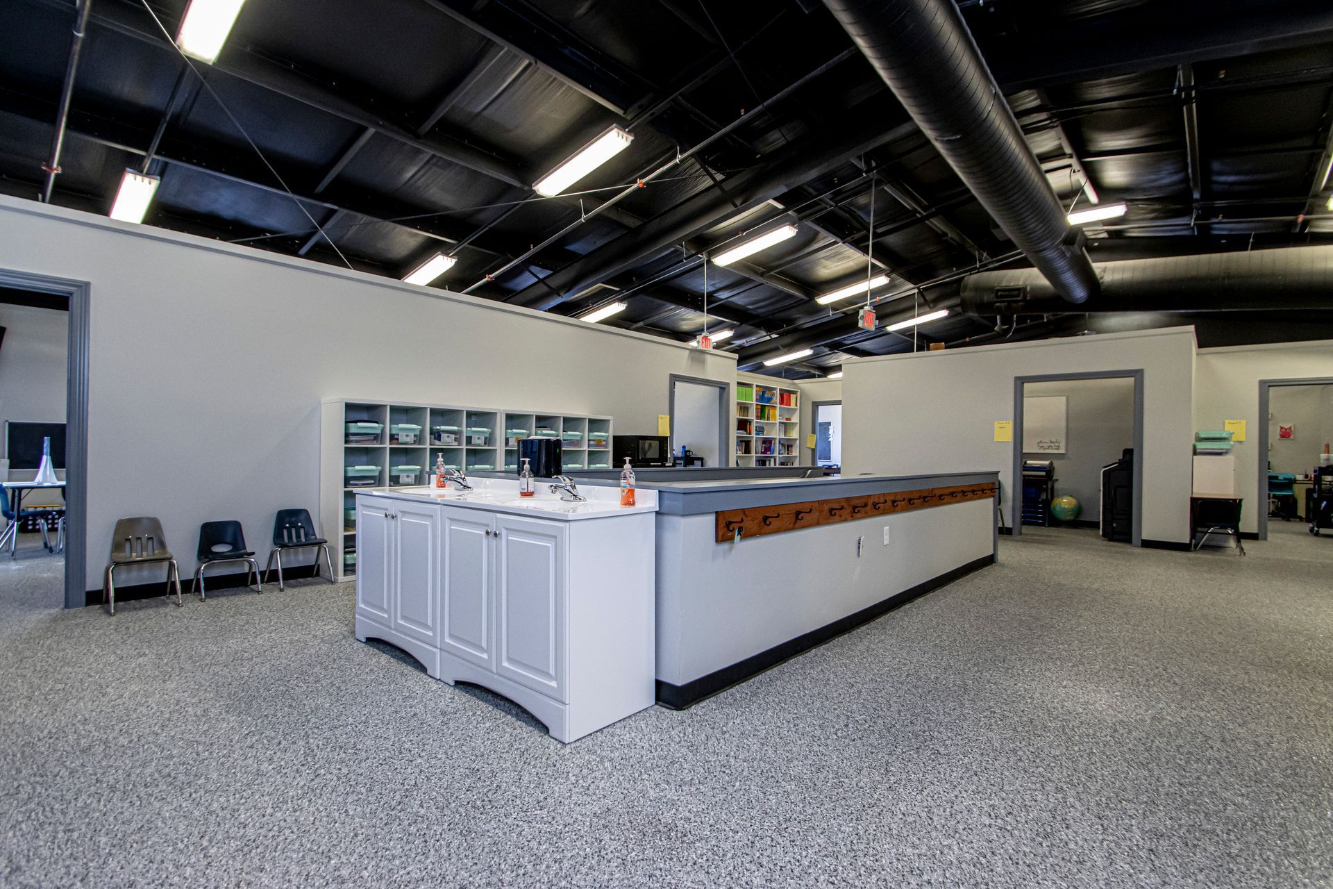 A wide-angle view of a modern classroom with grey cabinets, a long workstation, speckled flooring, and an open ceiling.