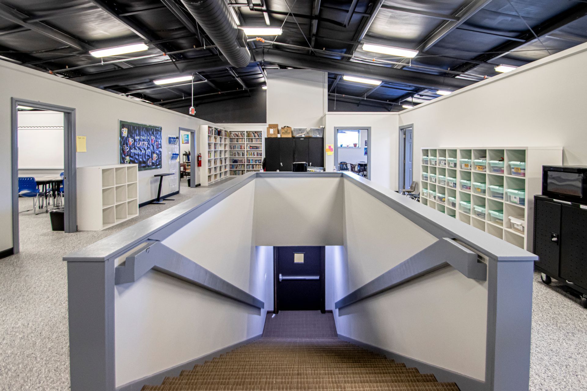 Staircase leading down in a modern, open-plan school hallway with white cubbies and exposed ceiling rafters.