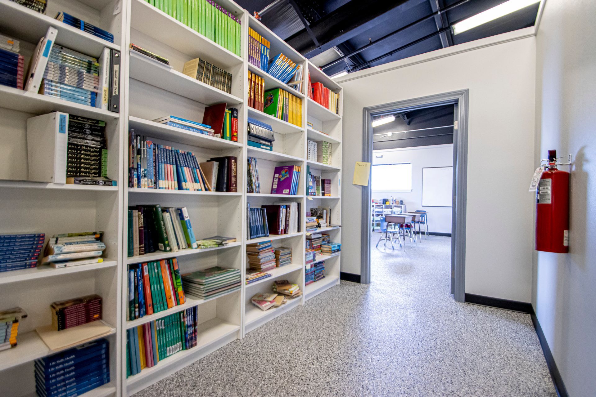 A hallway with tall, white bookshelves packed with books and a red fire extinguisher on the right wall.